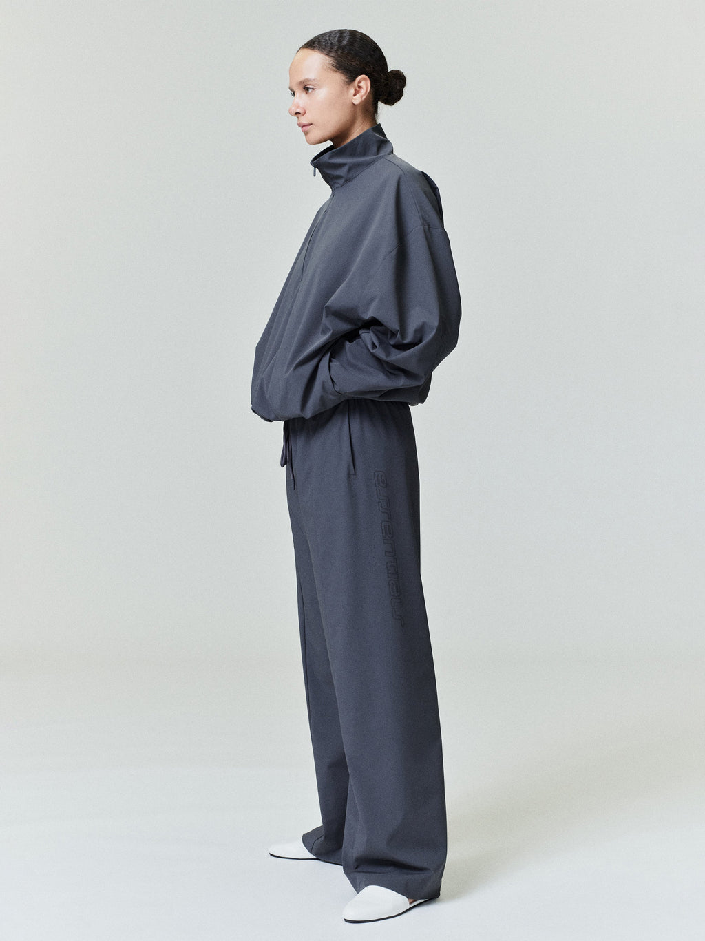 Woman in loose dark gray nylon half-zip track jacket and matching wide-leg pants standing in profile with hands in pockets