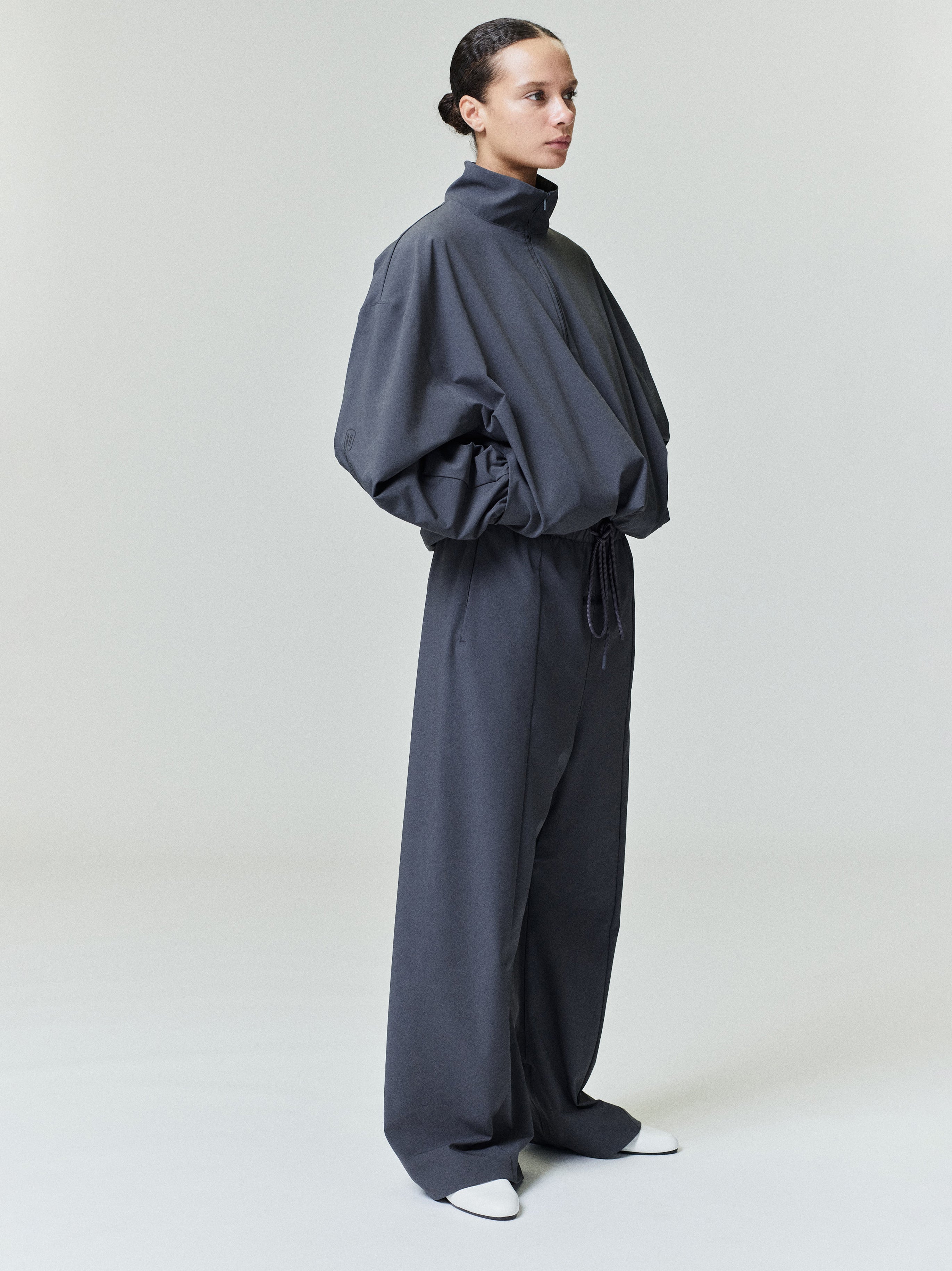 Woman wearing a dark gray oversized nylon half-zip track jacket with matching wide-leg pants and white shoes
