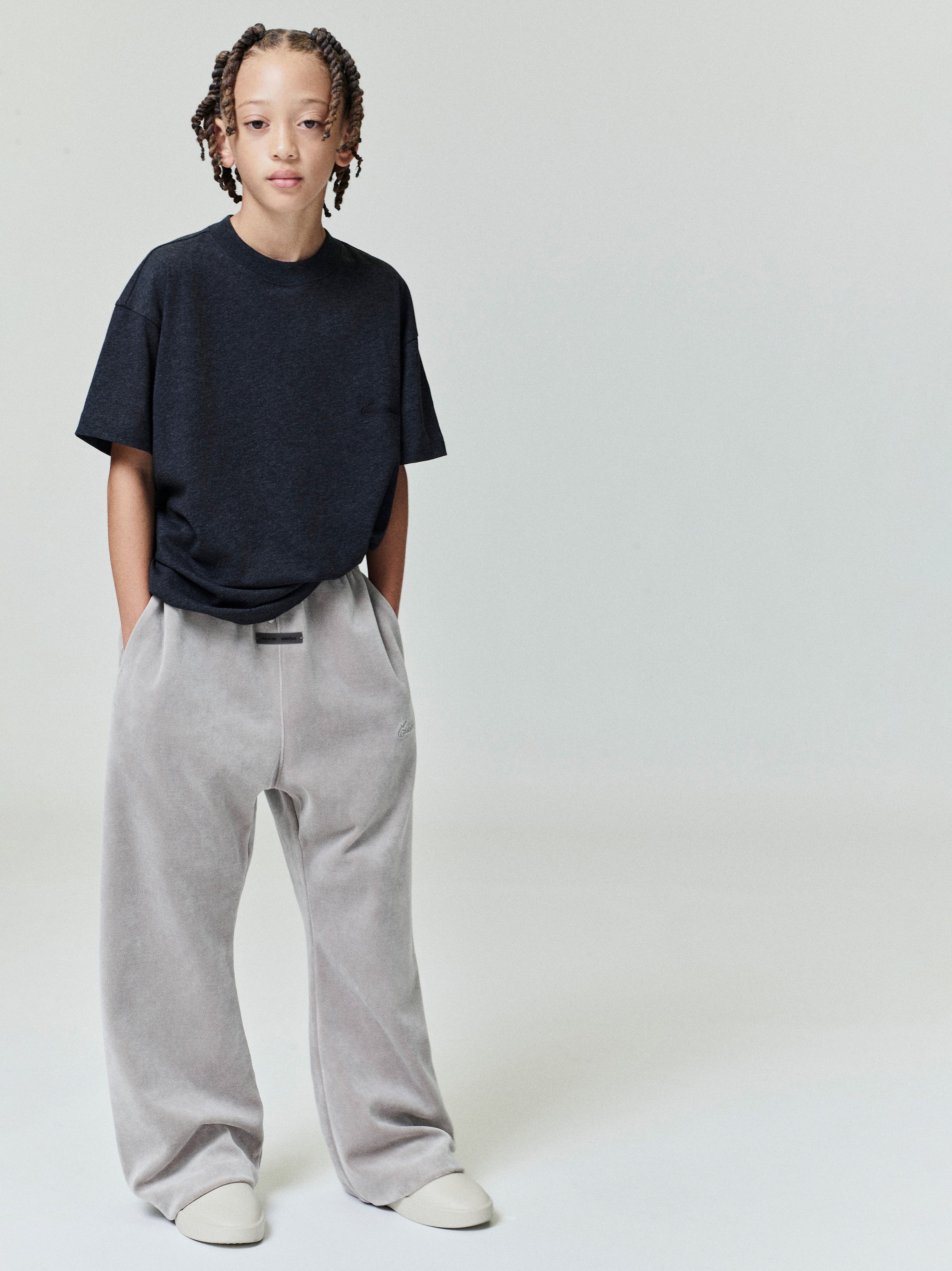 Full body of child wearing black Kids Embroidered Essential Tee with hands in pockets, standing against plain background, gallery position 0