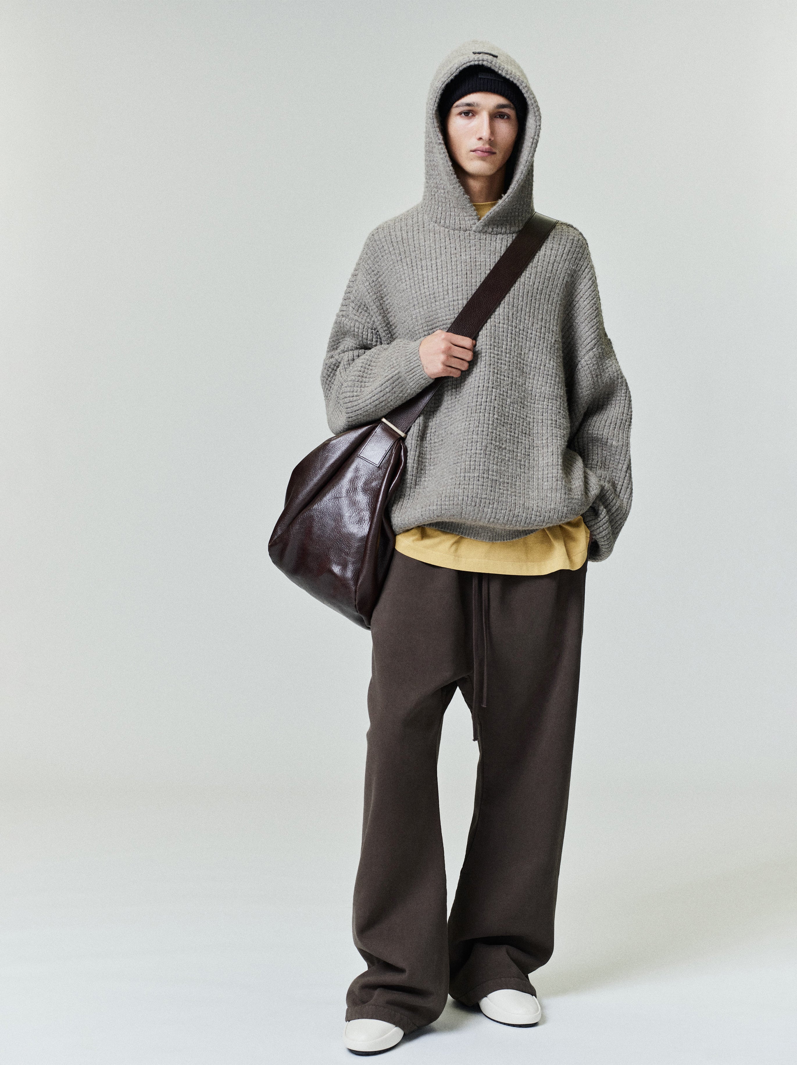 Full-length image of model wearing grey Heavy Waffle 90s Hoodie with hood up and oversized fit holding a dark leather shoulder bag