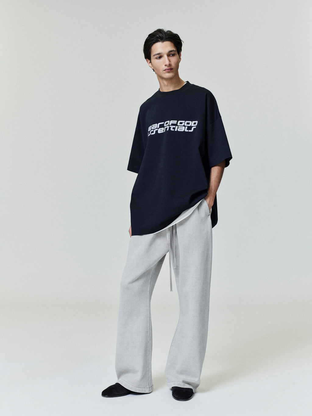 Male model wearing oversized black Ponte 90s Tee with white distorted logo, styled with grey wide-leg pants, gallery image 0005_HO25_F_L56_P07_2493.jpg