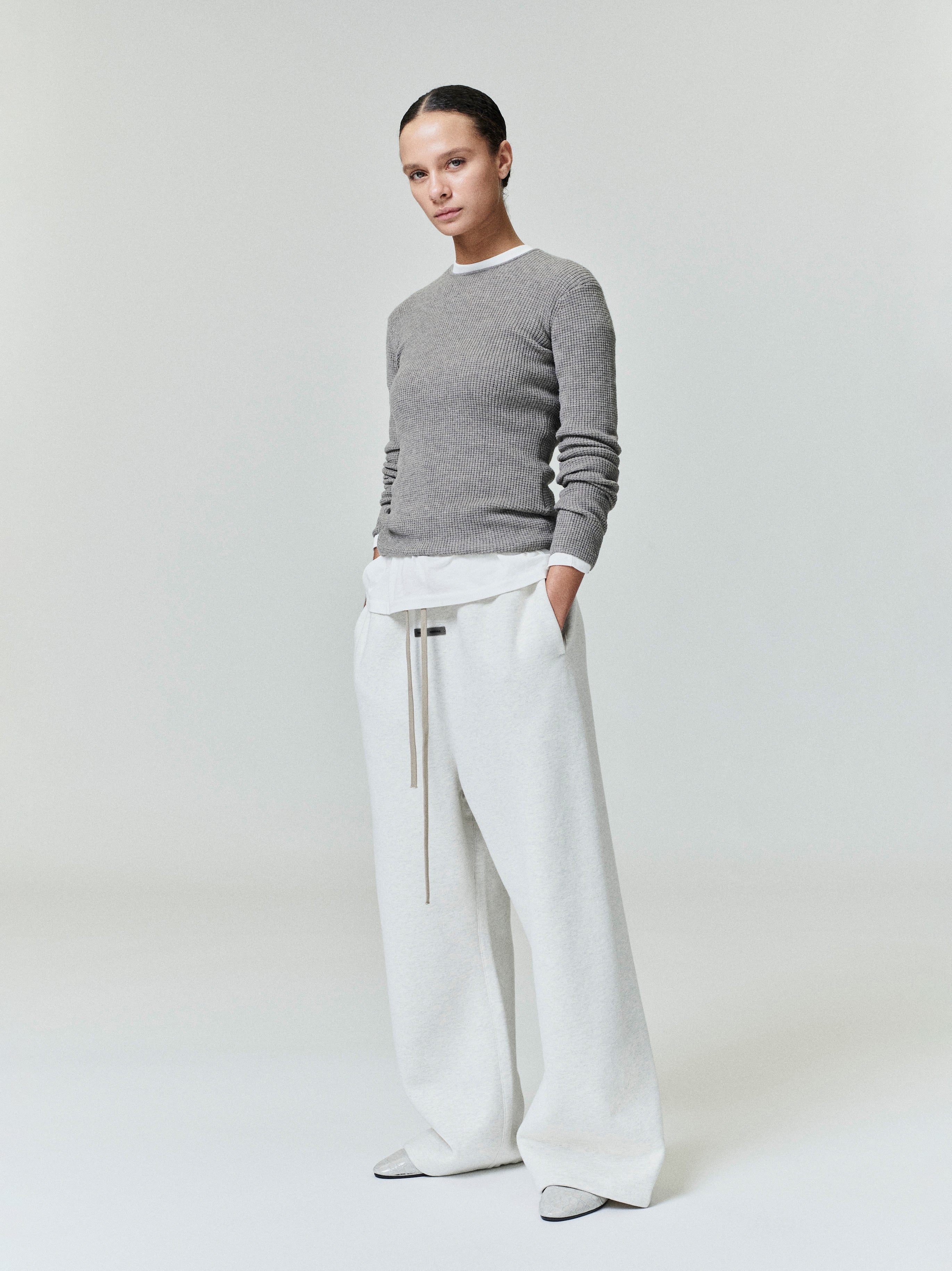 Woman wearing grey waffle textured long sleeve top and loose white wide leg pants standing against light background