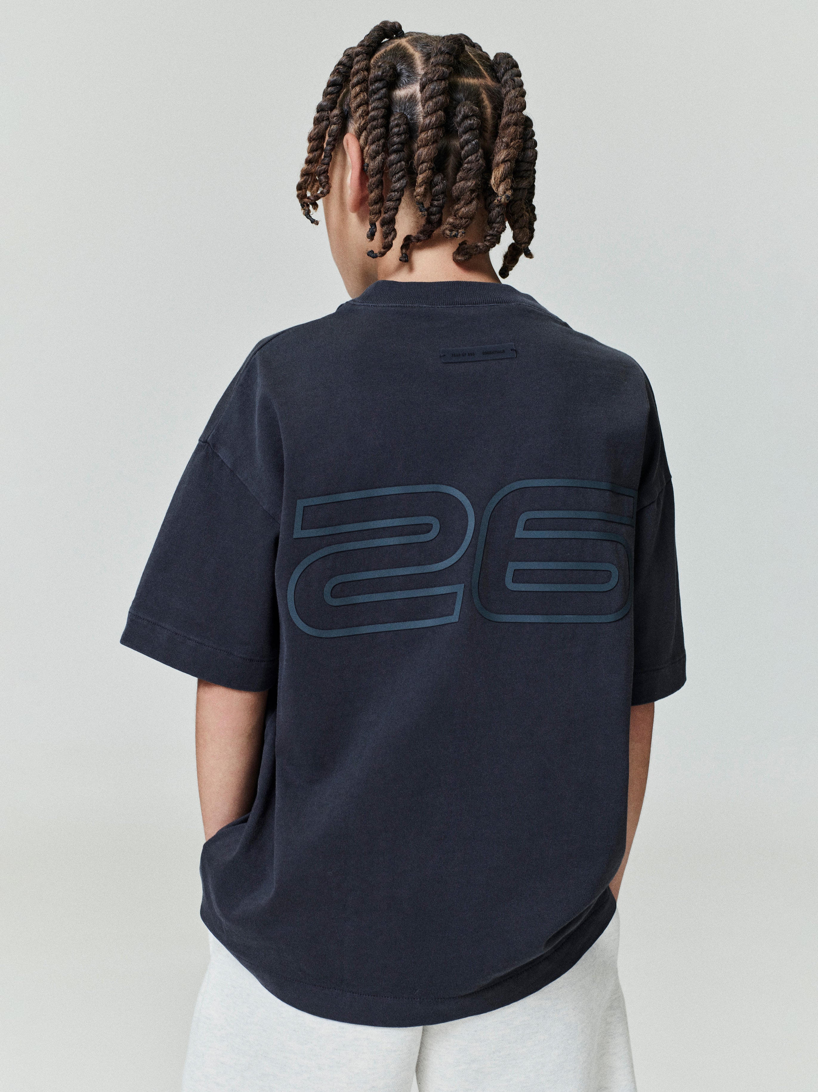 Back view of child wearing navy Kids Classic Short Sleeve Tee with large outlined "26" graphic, gallery position 0, file 0006_HO25_K_L75_4017.jpg