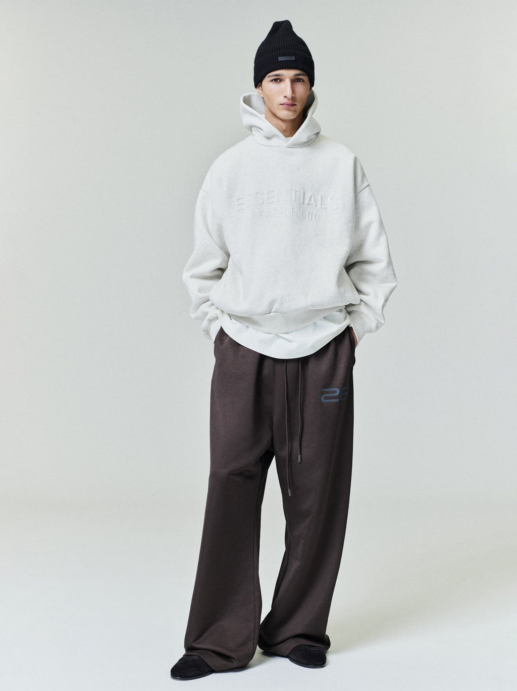 Model wearing oatmeal Embossed Essentials Classic Hoodie paired with wide-leg dark brown pants and black beanie, full body shot