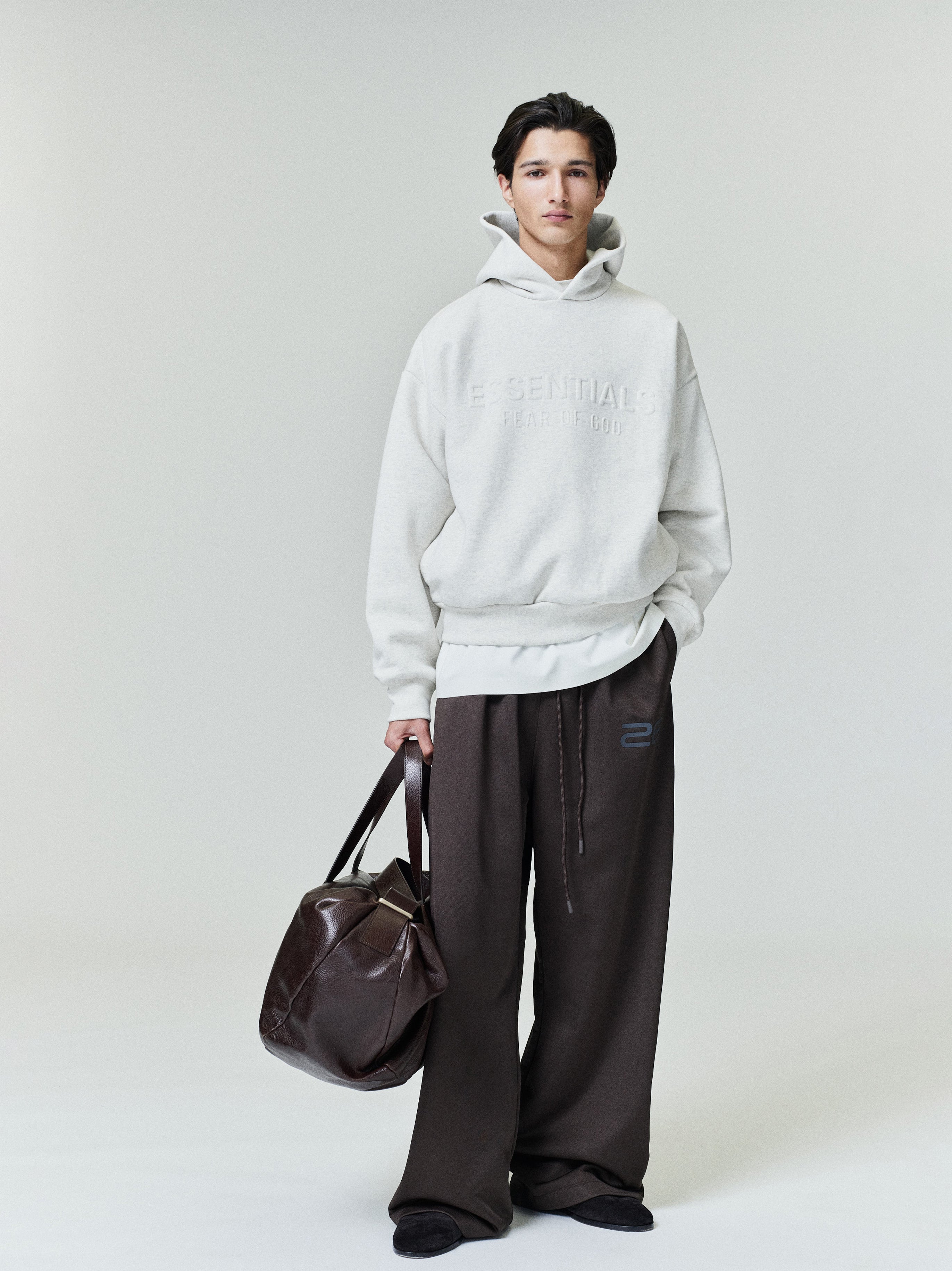 Model wearing oatmeal Embossed Essentials Classic Hoodie standing with hand in pocket and holding dark brown leather bag, studio shot