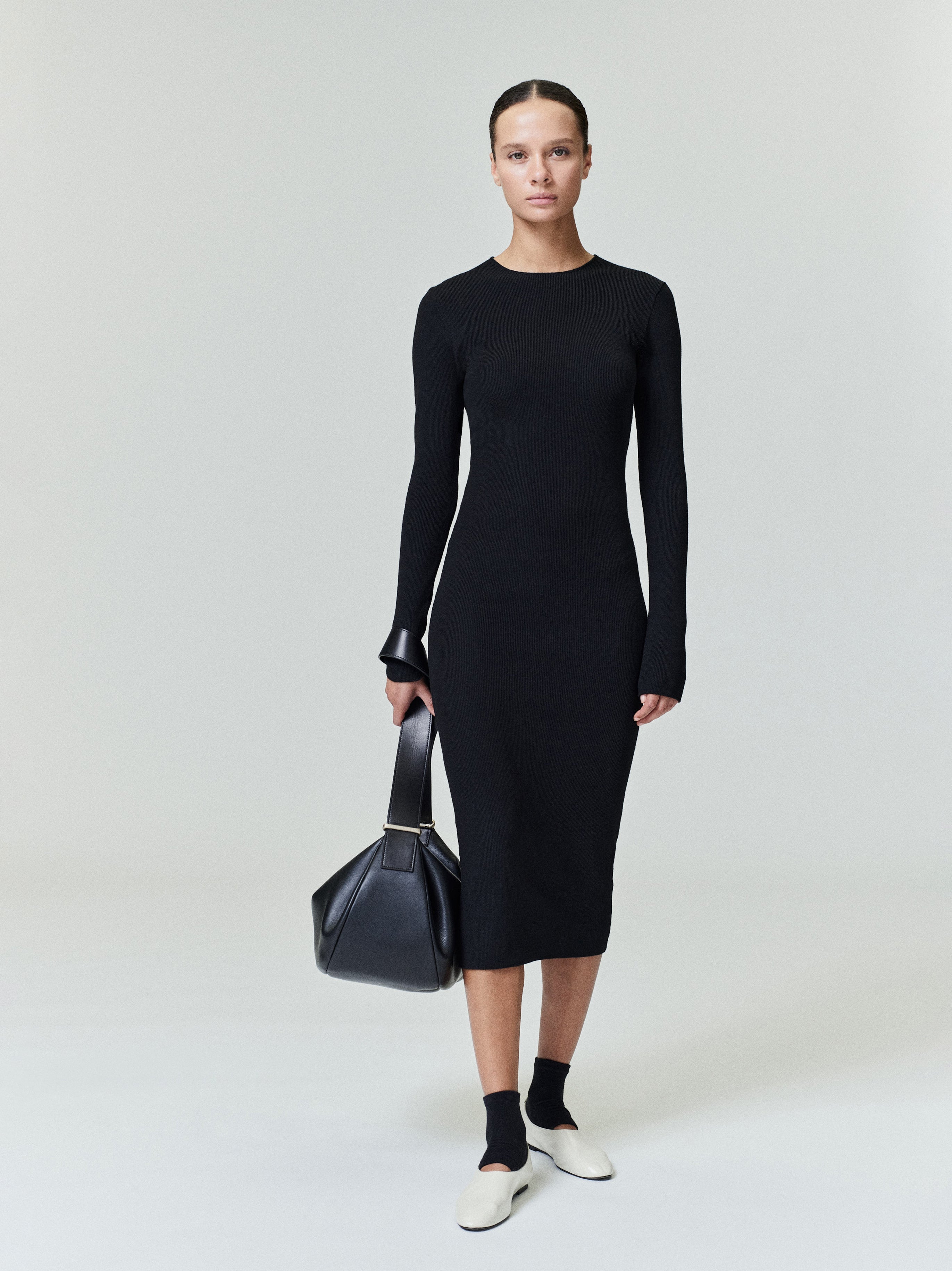 Model wearing black fitted ribbed long sleeve midi dress with rounded neckline holding large black leather handbag