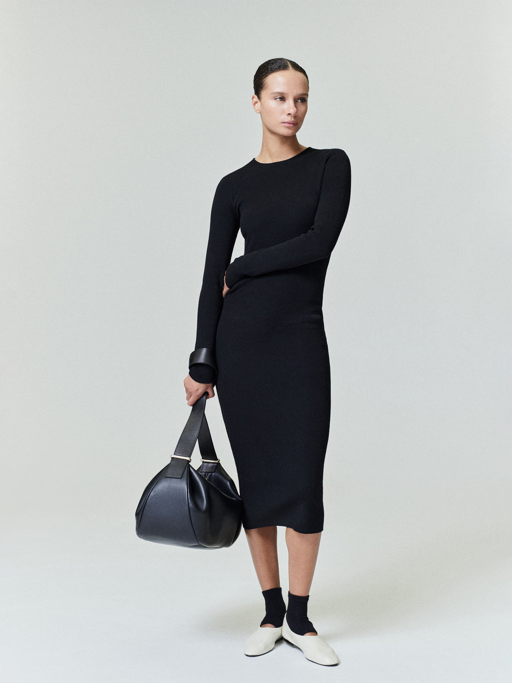 Woman in black ribbed long sleeve midi dress holding black leather bag against gray background