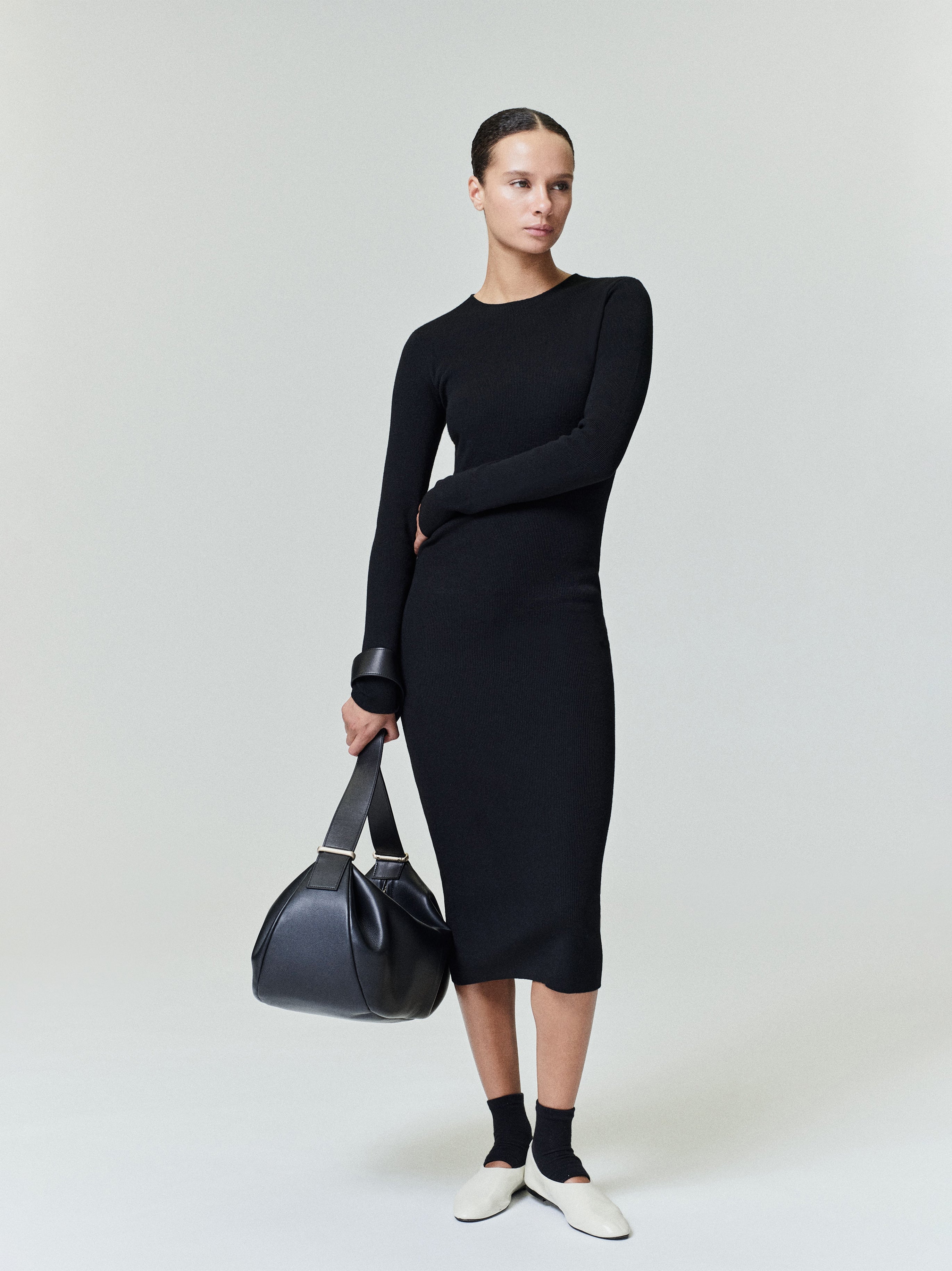 Woman in black ribbed long sleeve midi dress holding black leather bag against gray background