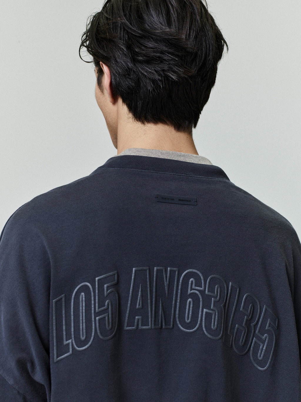Rear view of man wearing dark Los Angeles 90s Tee with large embossed numeric lettering on back neckline