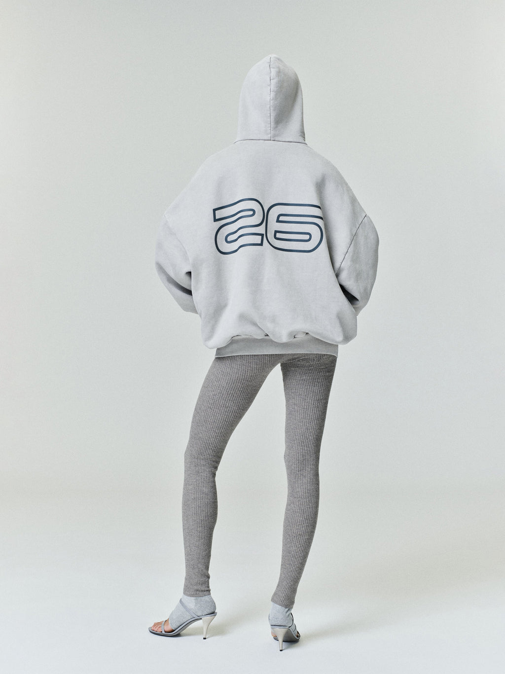 Model seen from behind wearing light gray Heavy Fleece 90s Hoodie with outlined number 26 on back, paired with ribbed leggings and heels