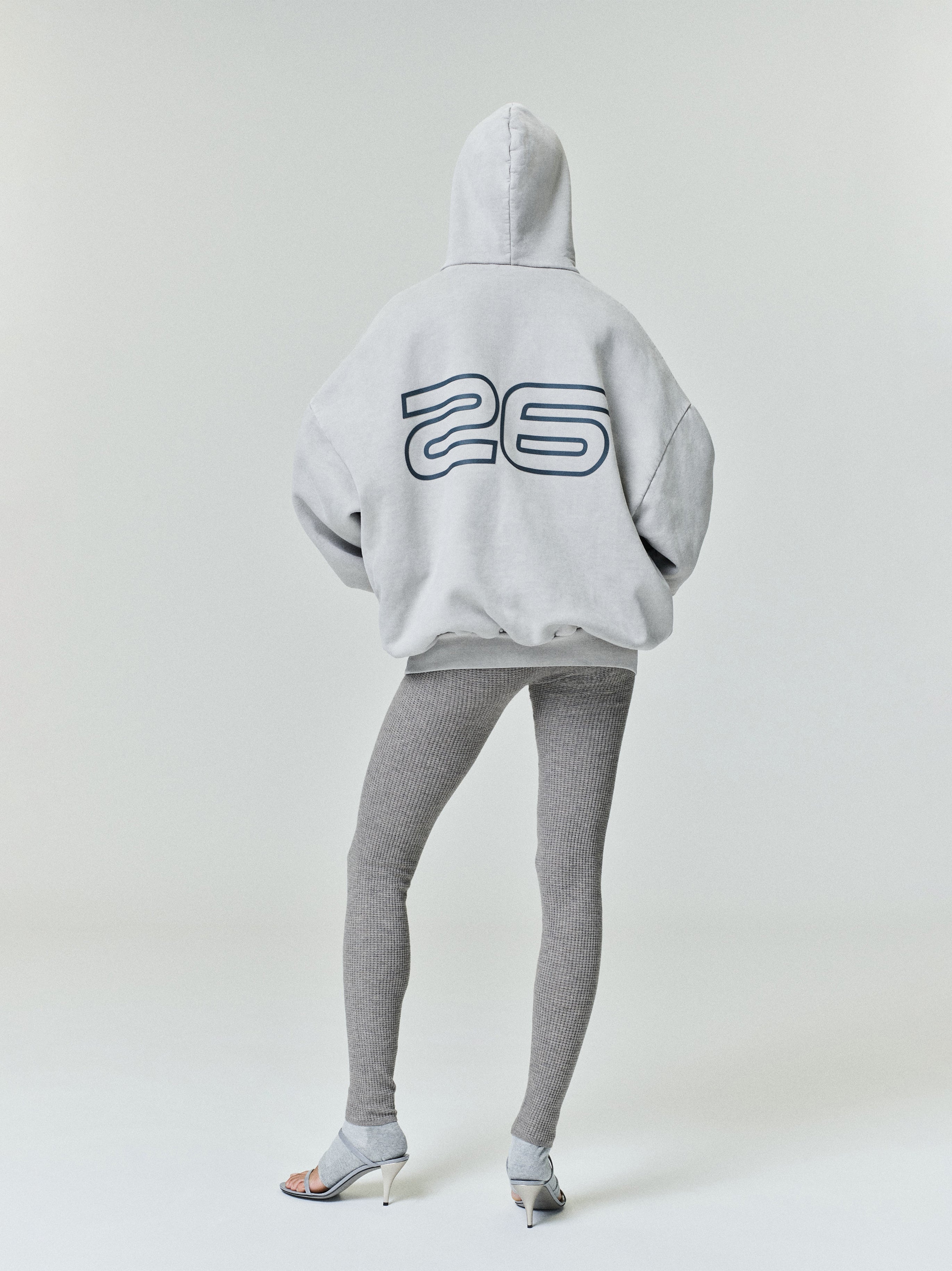 Model seen from behind wearing light gray Heavy Fleece 90s Hoodie with outlined number 26 on back, paired with ribbed leggings and heels