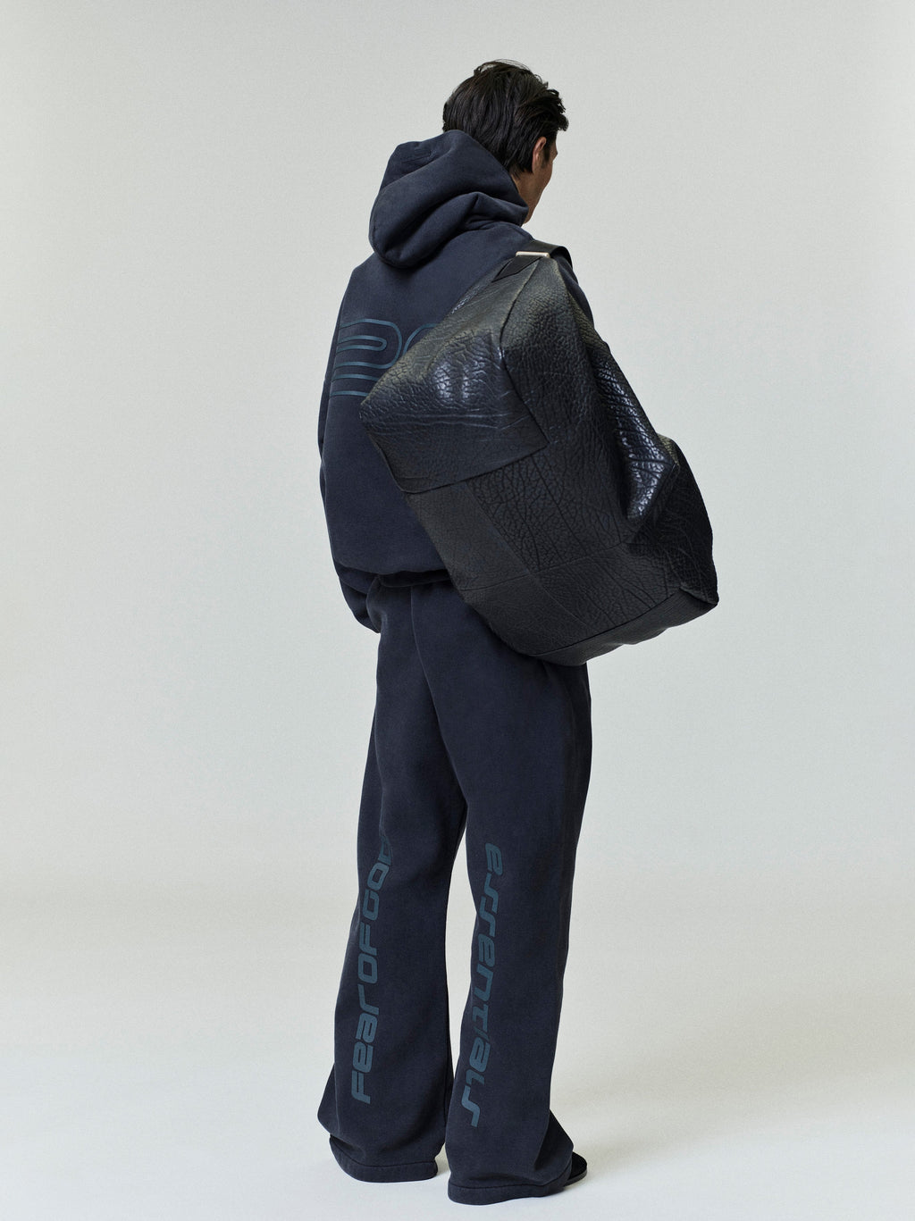 Rear view of model wearing dark navy Heavy Fleece 90s Hoodie and matching pants carrying oversized black textured bag gallery 0