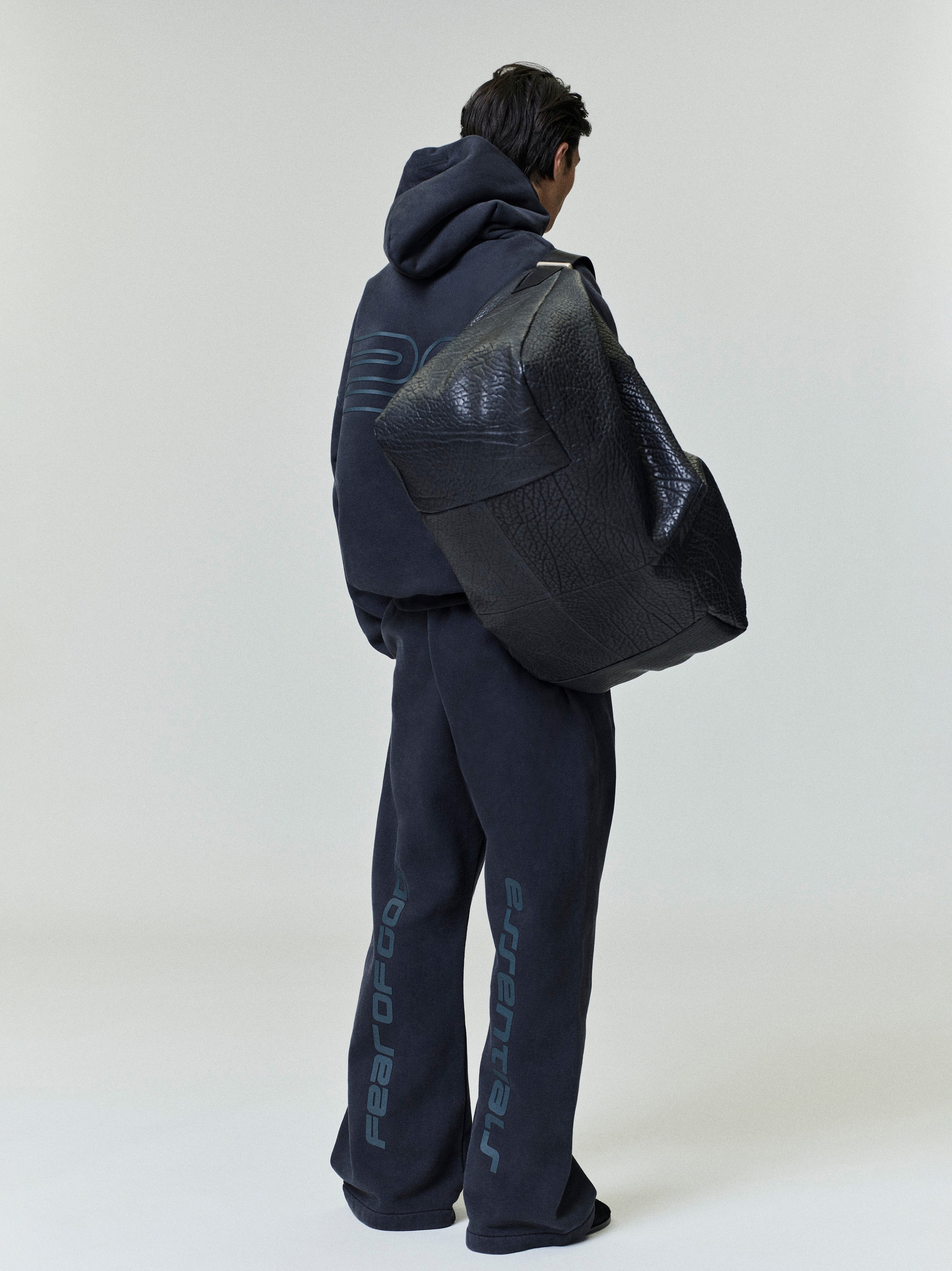 Rear view of model wearing dark navy Heavy Fleece 90s Hoodie and matching pants carrying oversized black textured bag gallery 0