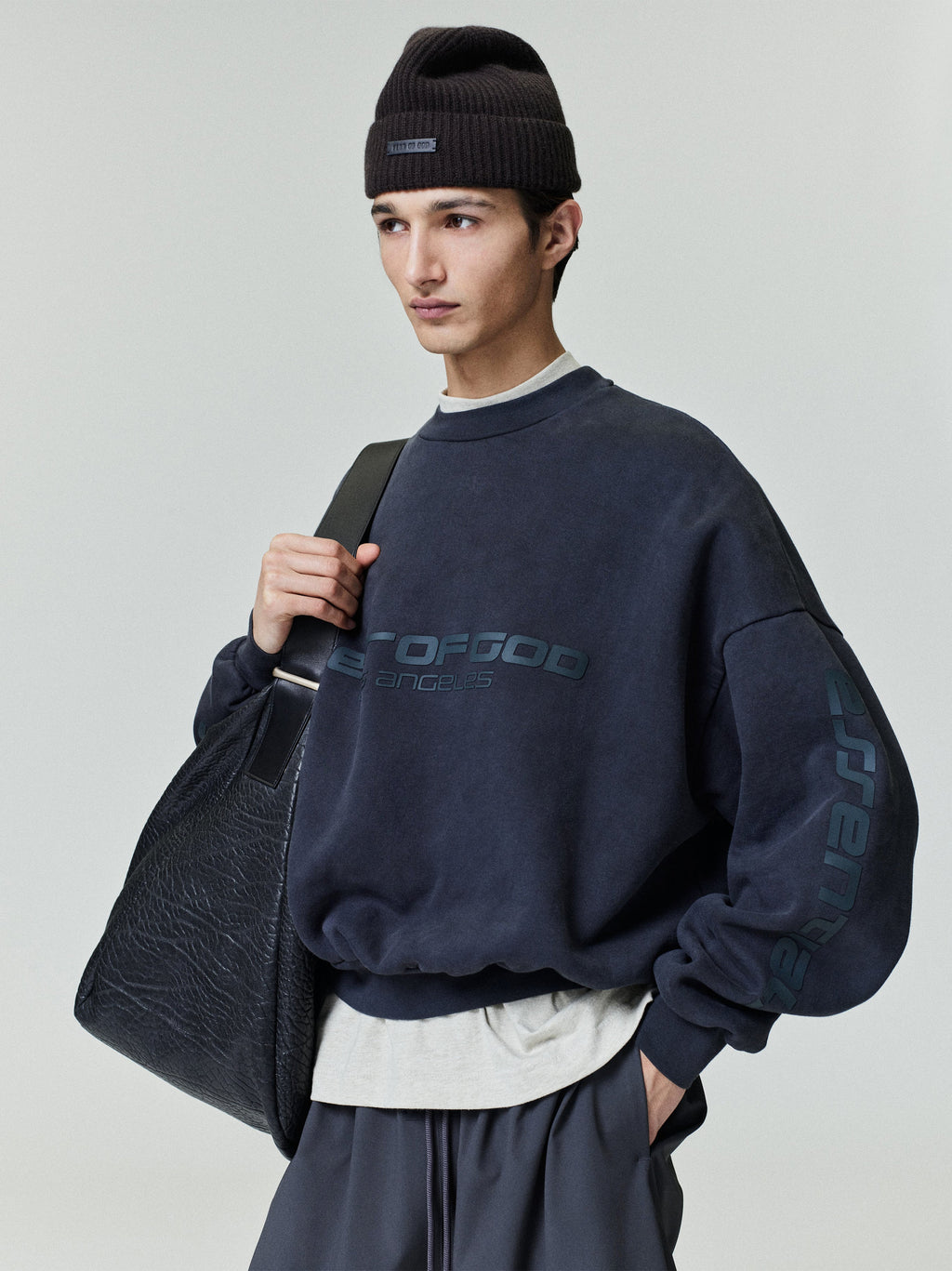 Male model wearing navy Los Angeles heavy fleece cropped sweatshirt with black beanie and large textured leather tote bag