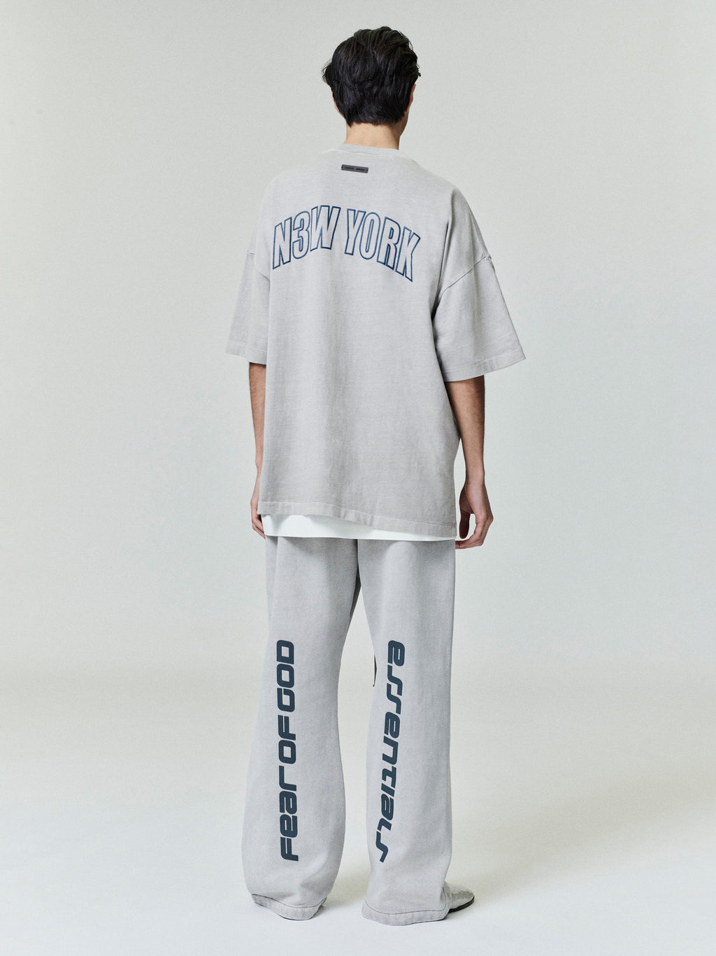 Back view of oversized light grey New York 90s Tee with curved navy print paired with wide-leg pants featuring vertical text designs