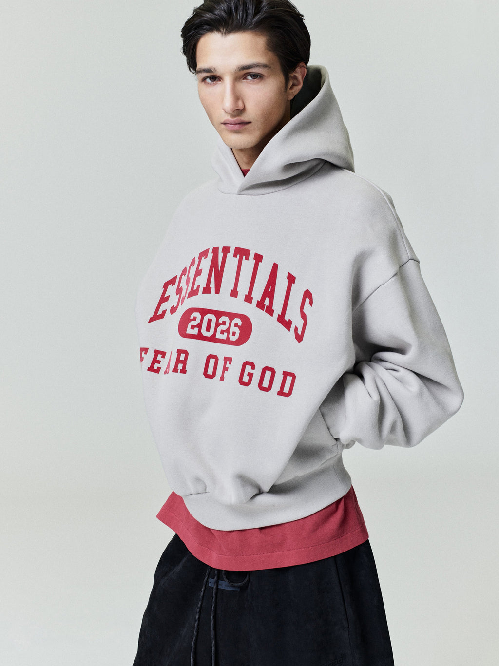 Side view of model wearing light grey Classic Hoodie with red Essentials 2026 text and layered red shirt underneath