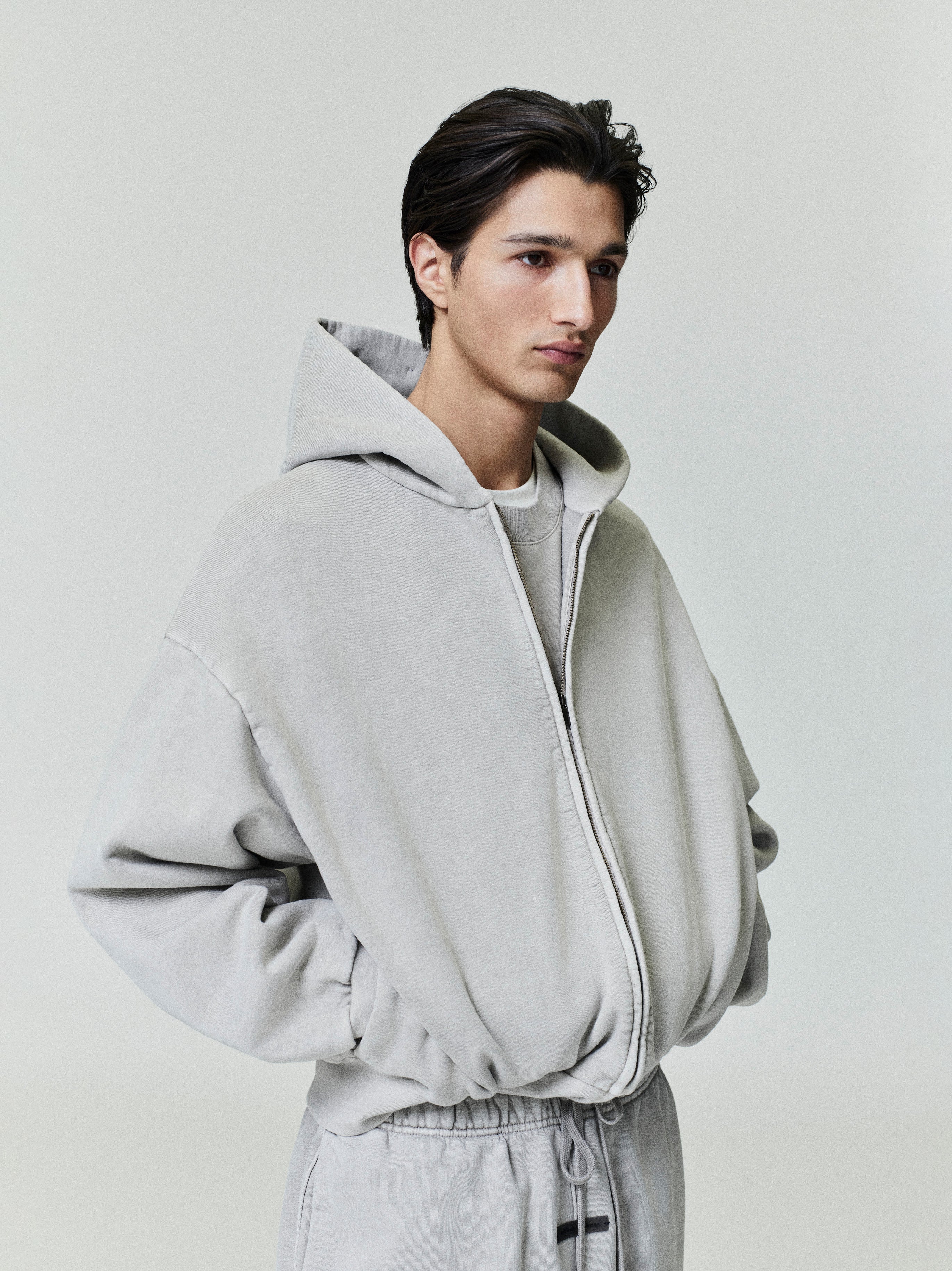 Side angle of light grey Embroidered Classic Full Zip Hoodie with oversized fit and model’s hands in pockets on neutral background