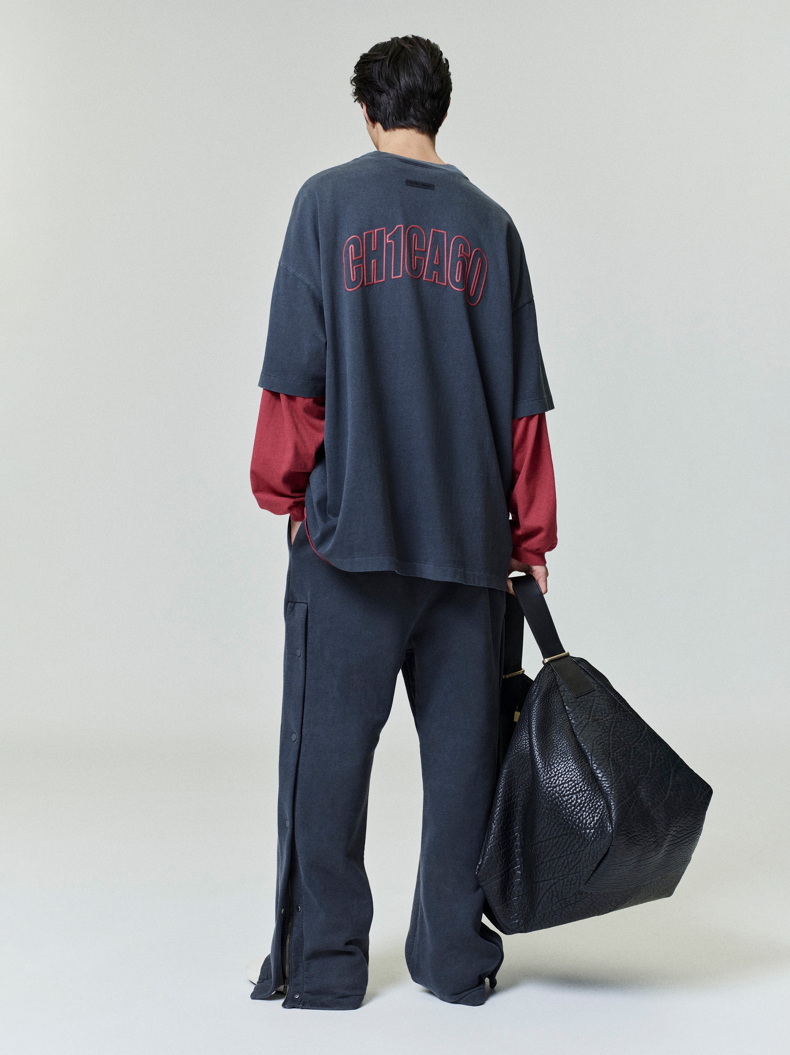 Rear view of model wearing oversized Chicago 90s Tee over red long sleeves, loose pants, and holding large black bag
