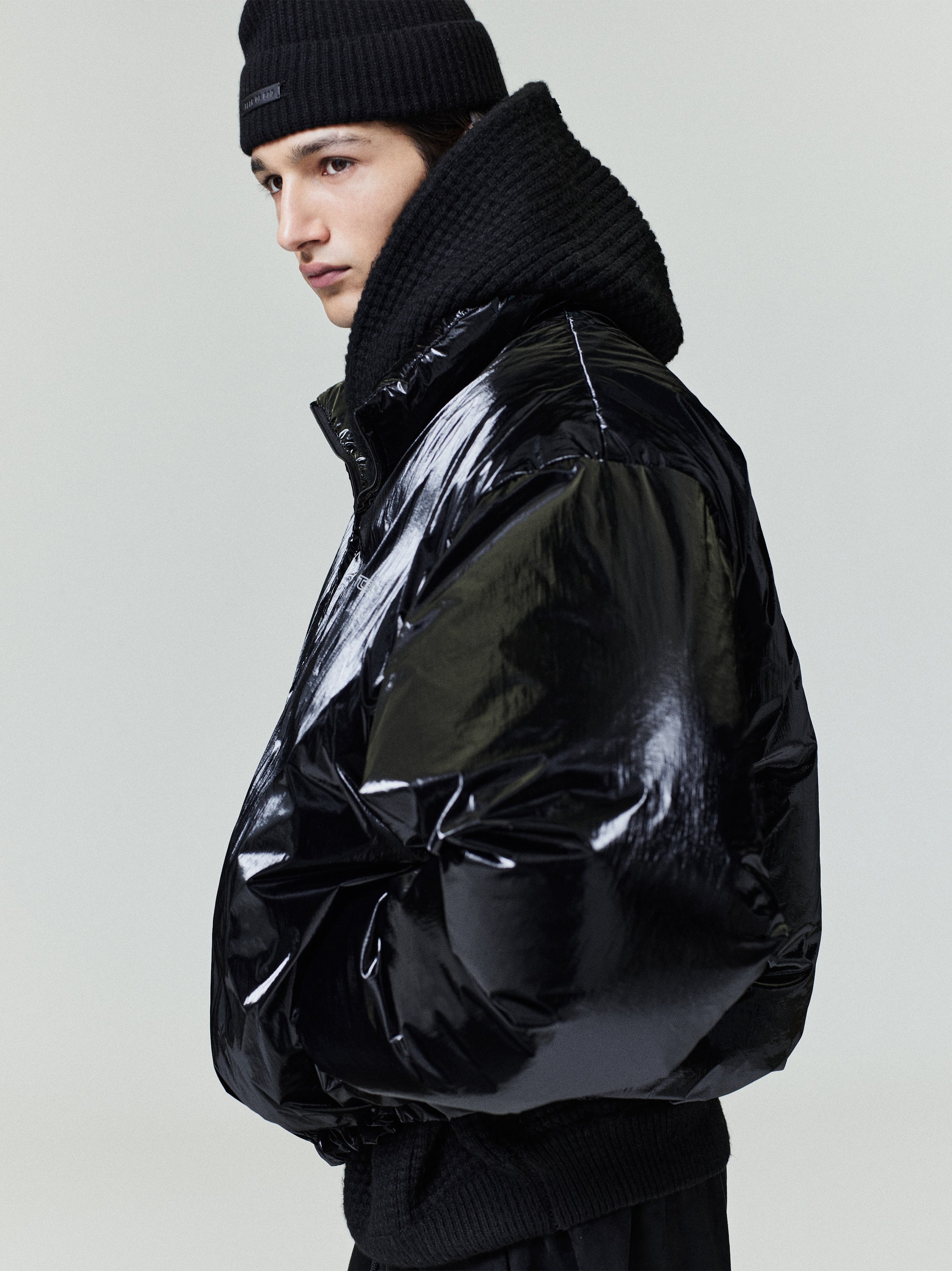 Profile of model wearing glossy black ripstop nylon puffer jacket with high collar and knit beanie, gallery position 0