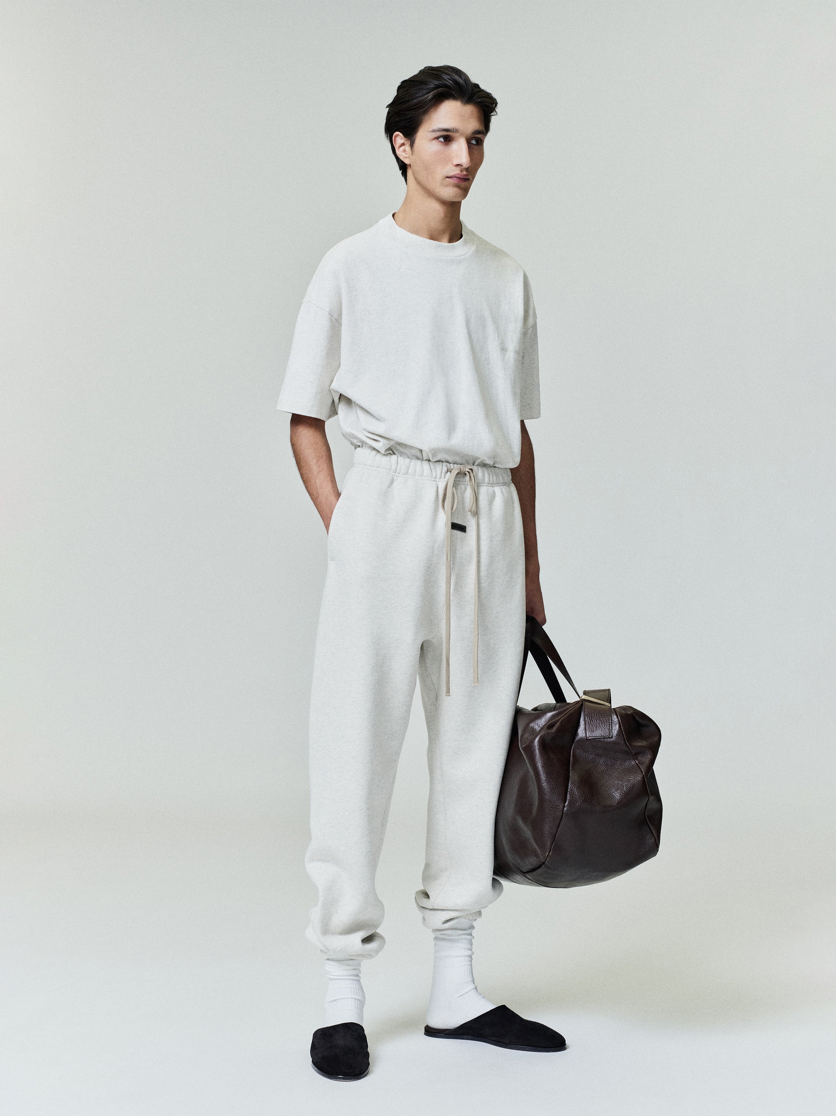 Model wearing off-white Classic Sweatpant with extended beige drawstring and relaxed fit holding dark brown leather bag against plain background