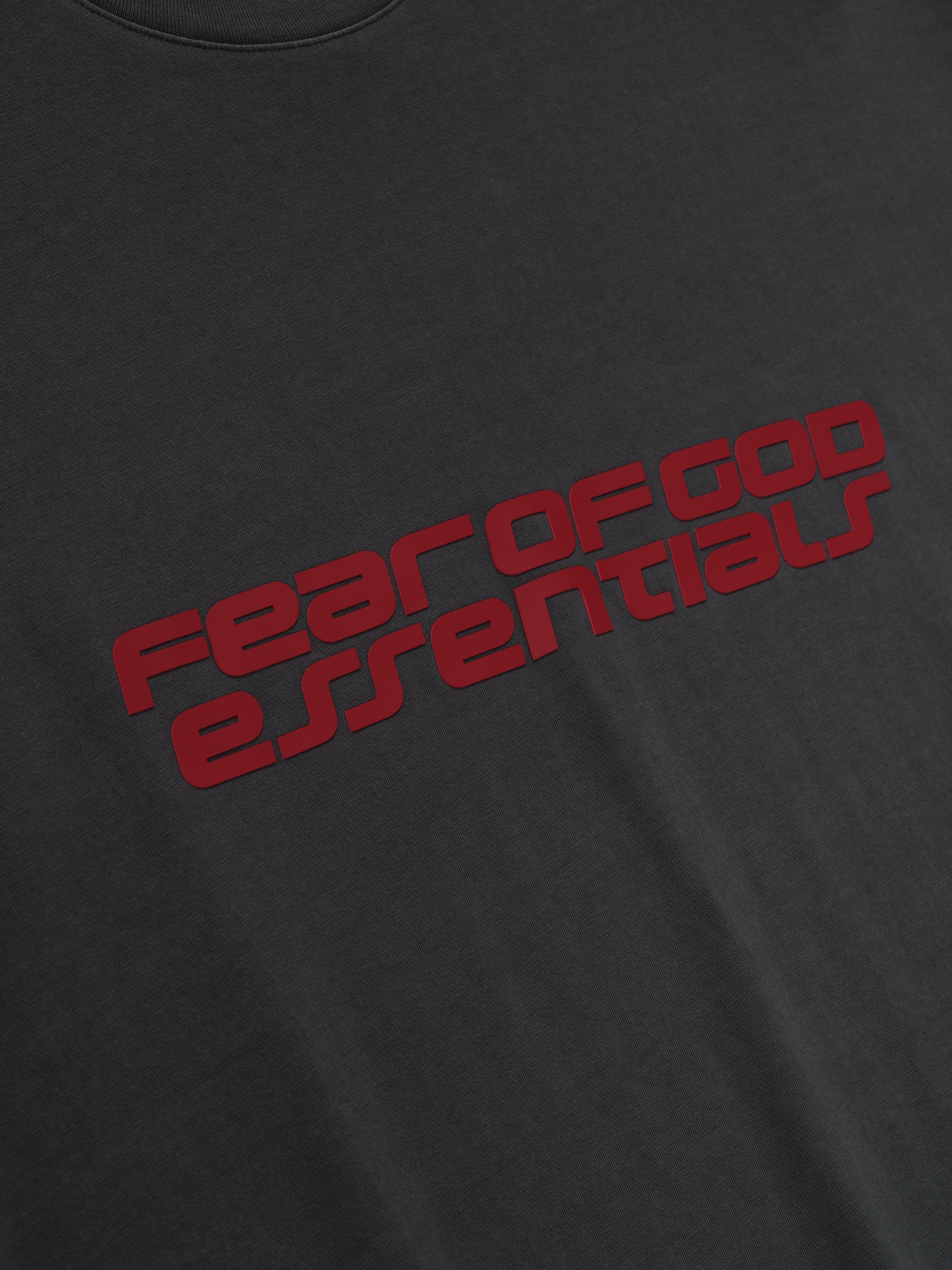 Close-up of iron grey Fear of God Essentials 90s Tee showing bold red logo text on smooth soft fabric front, gallery first image