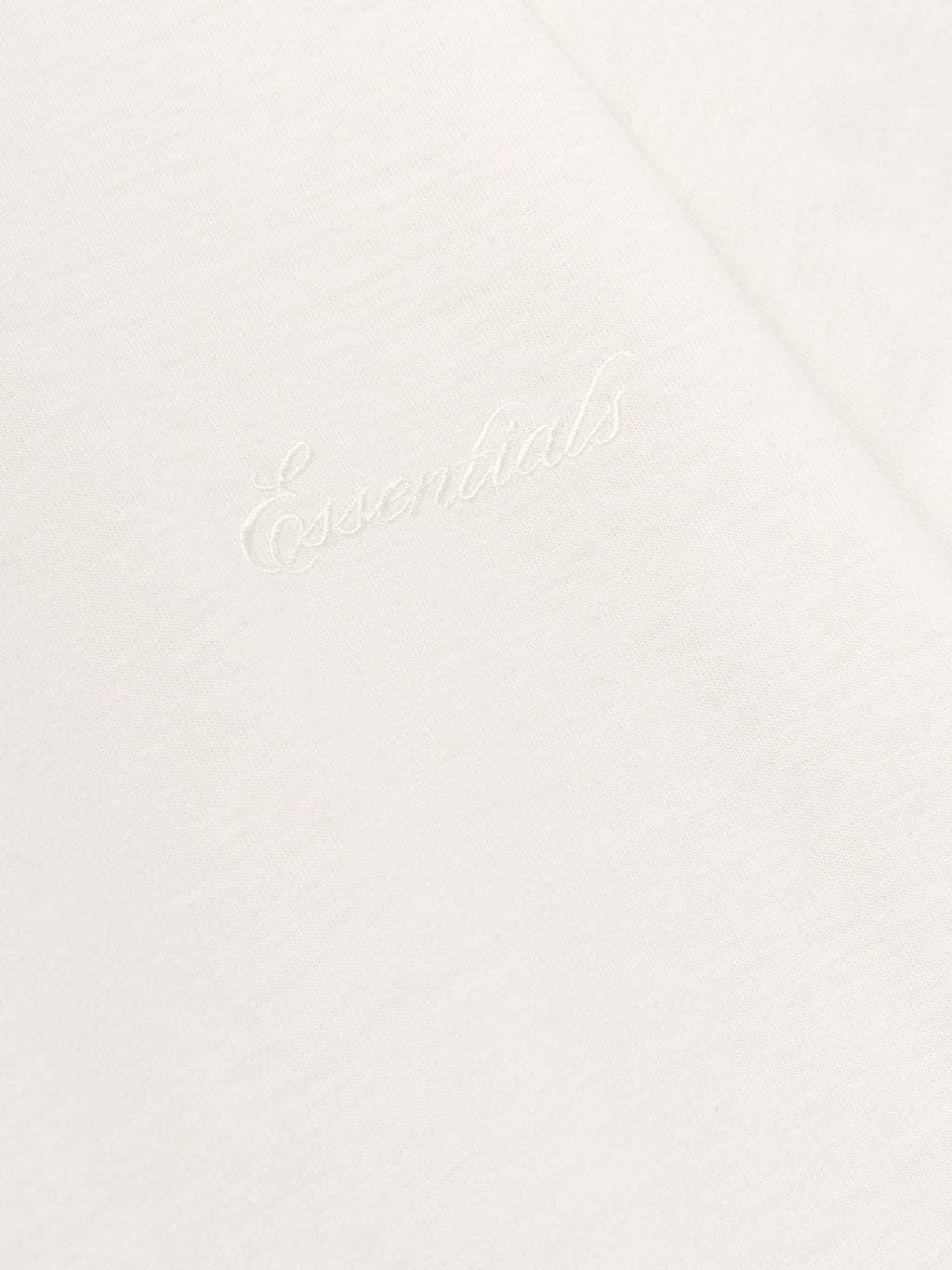 Close-up of ivory Womens Embroidered Classic Tee showing subtle "Essentials" chest embroidery on soft fabric backdrop
