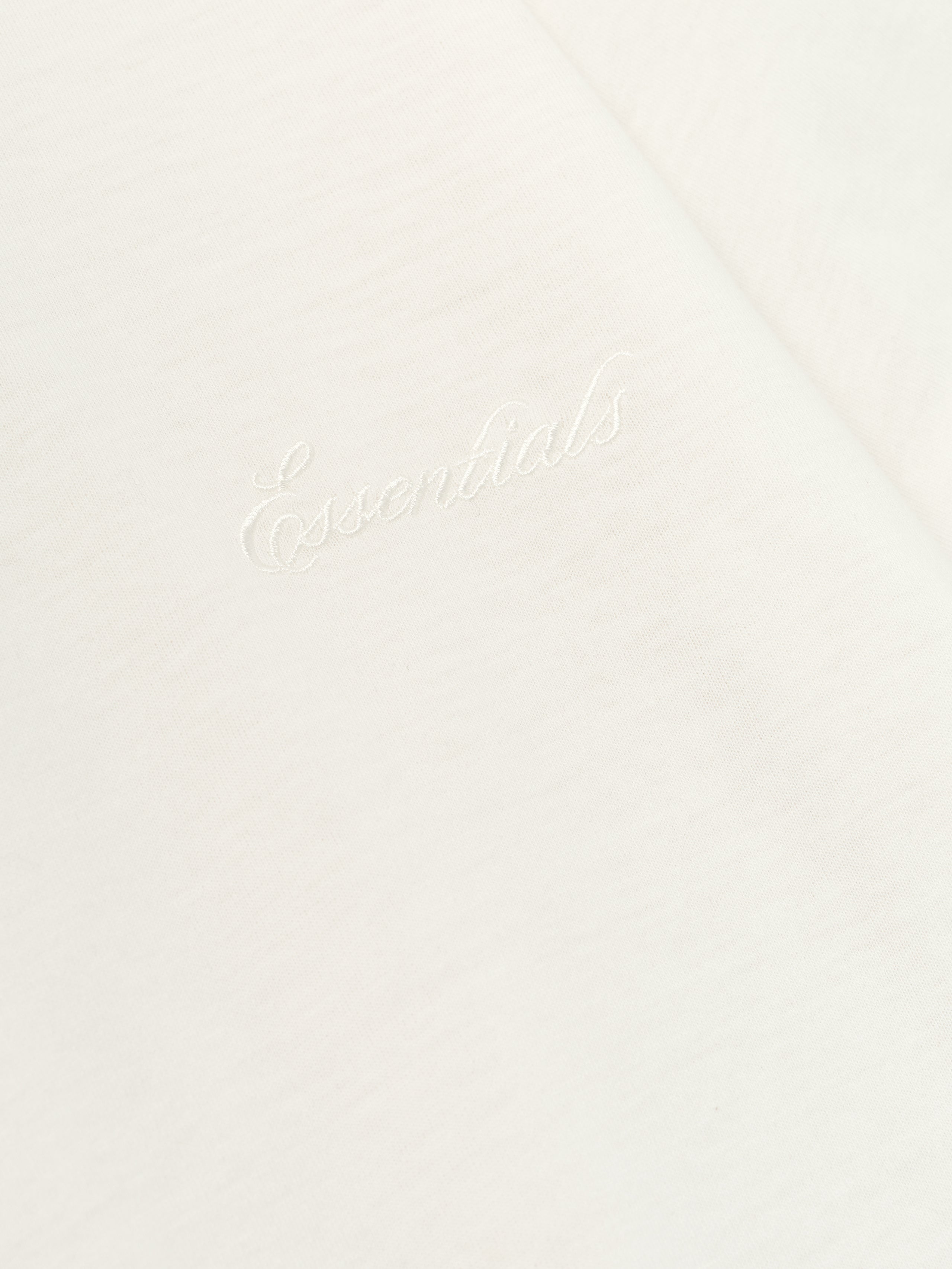 Close-up of ivory Womens Embroidered Classic Tee showing subtle "Essentials" chest embroidery on soft fabric backdrop