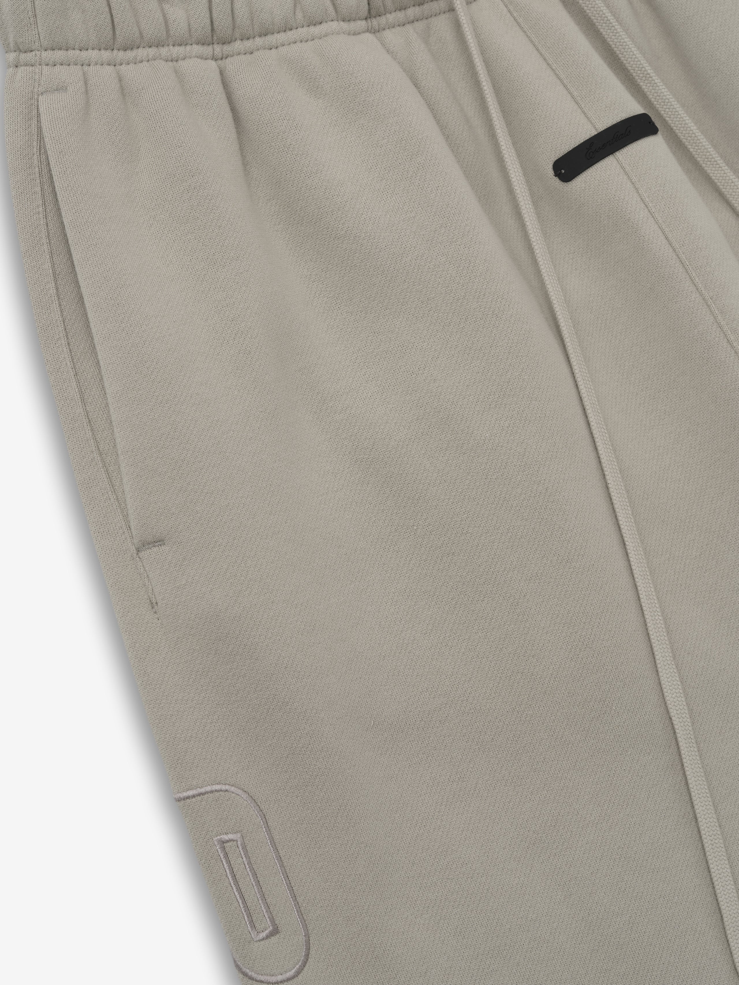 close-up of light taupe relaxed fit sweatpants featuring elastic waistband, drawstring, and tonal embroidered logo detail on leg
