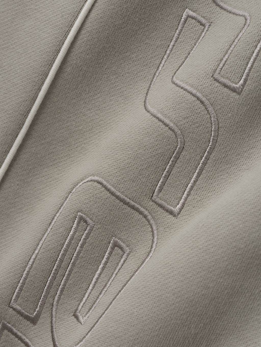 close-up of light taupe fabric with tonal embroidered letter detail and white sportswear piping on relaxed sweatpant