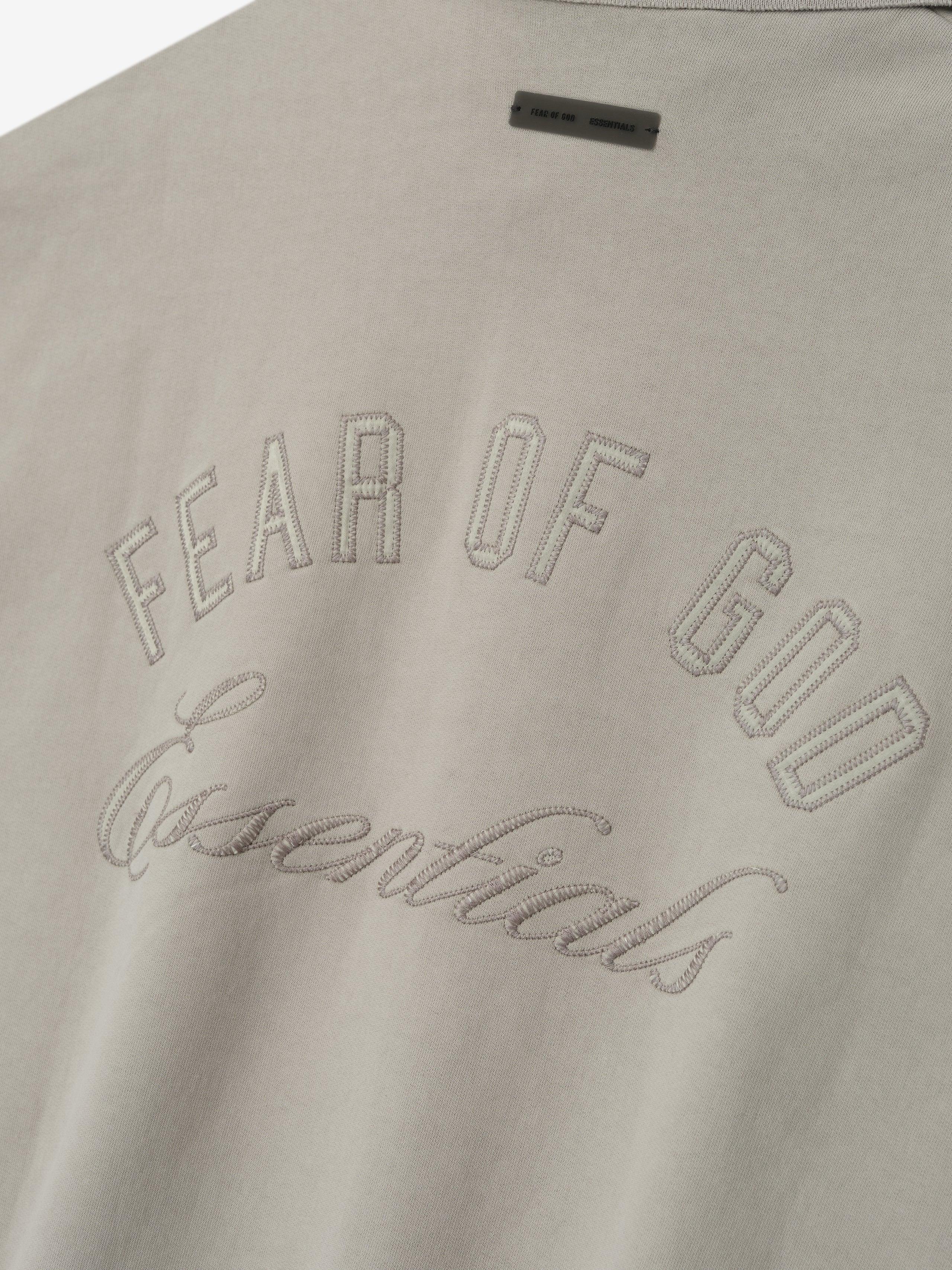 Close-up of smoke grey fabric with stitched "Fear of God Essentials" text and small rectangular label above on sweatshirt back.