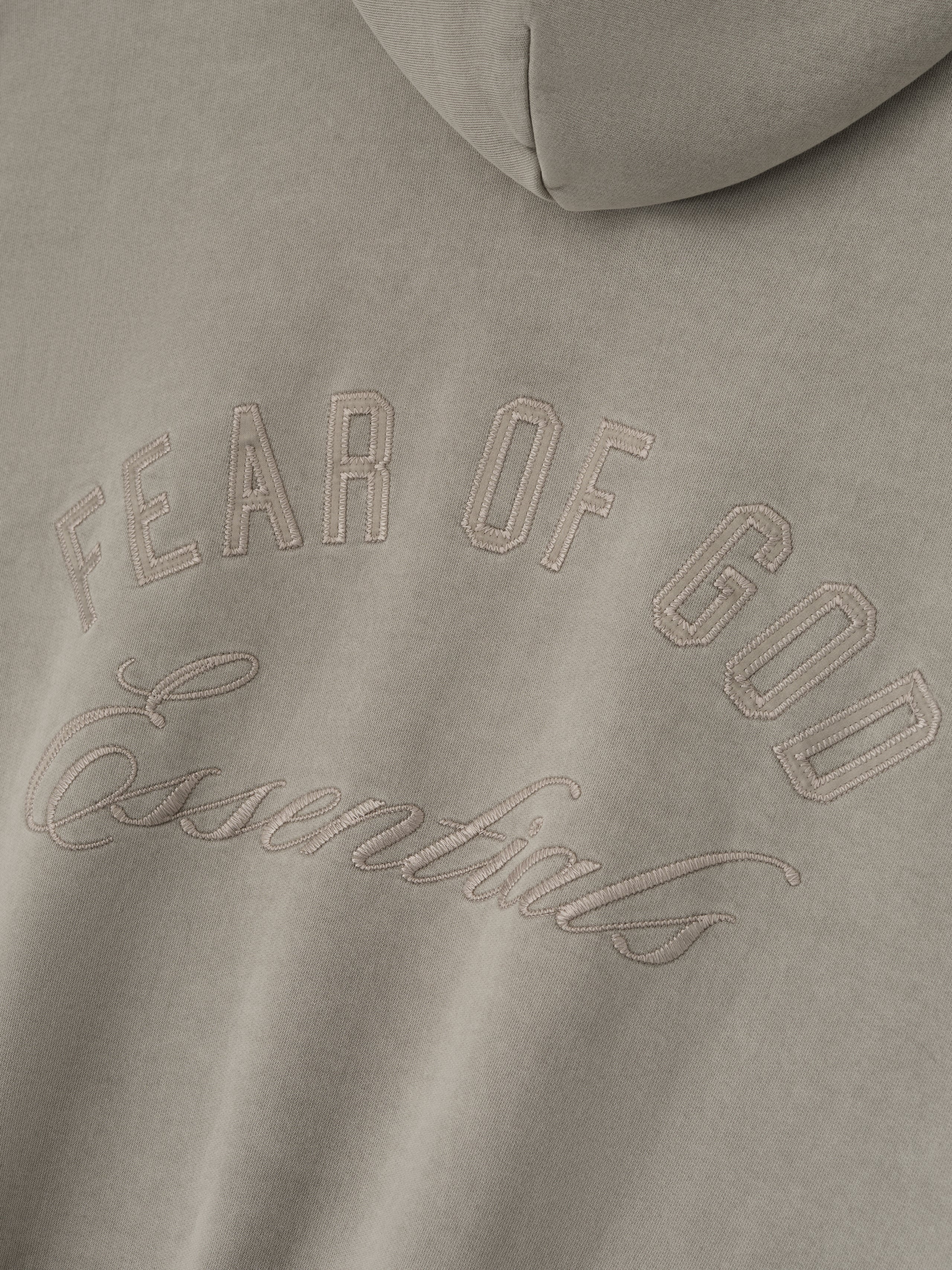 Close-up of smoke grey Embroidered Classic Full Zip Hoodie showing tonal Fear of God Essentials logo on hood section