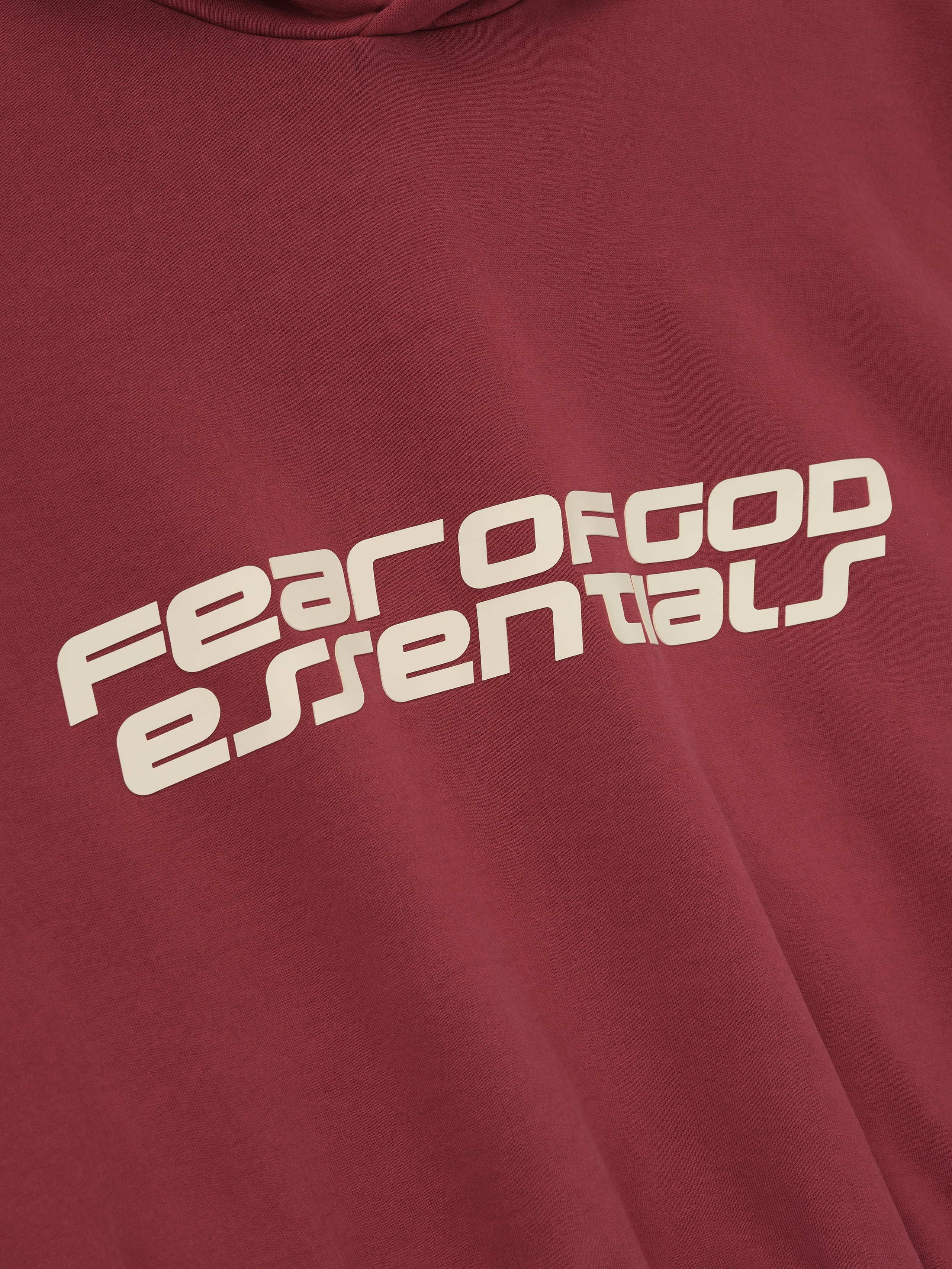 Close-up of crimson cropped relaxed hoodie featuring large slanted Fear of God Essentials logo in cream on front