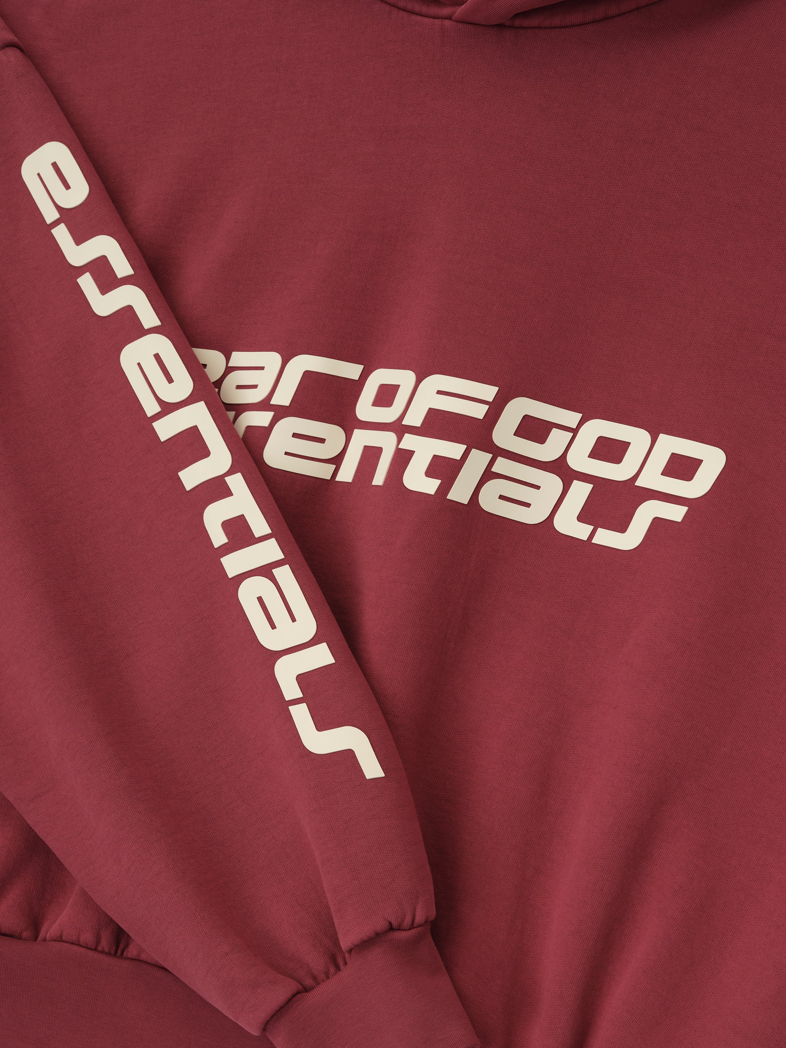Close-up of crimson cropped relaxed hoodie showing white Fear of God Essentials logo on chest and printed sleeve detail