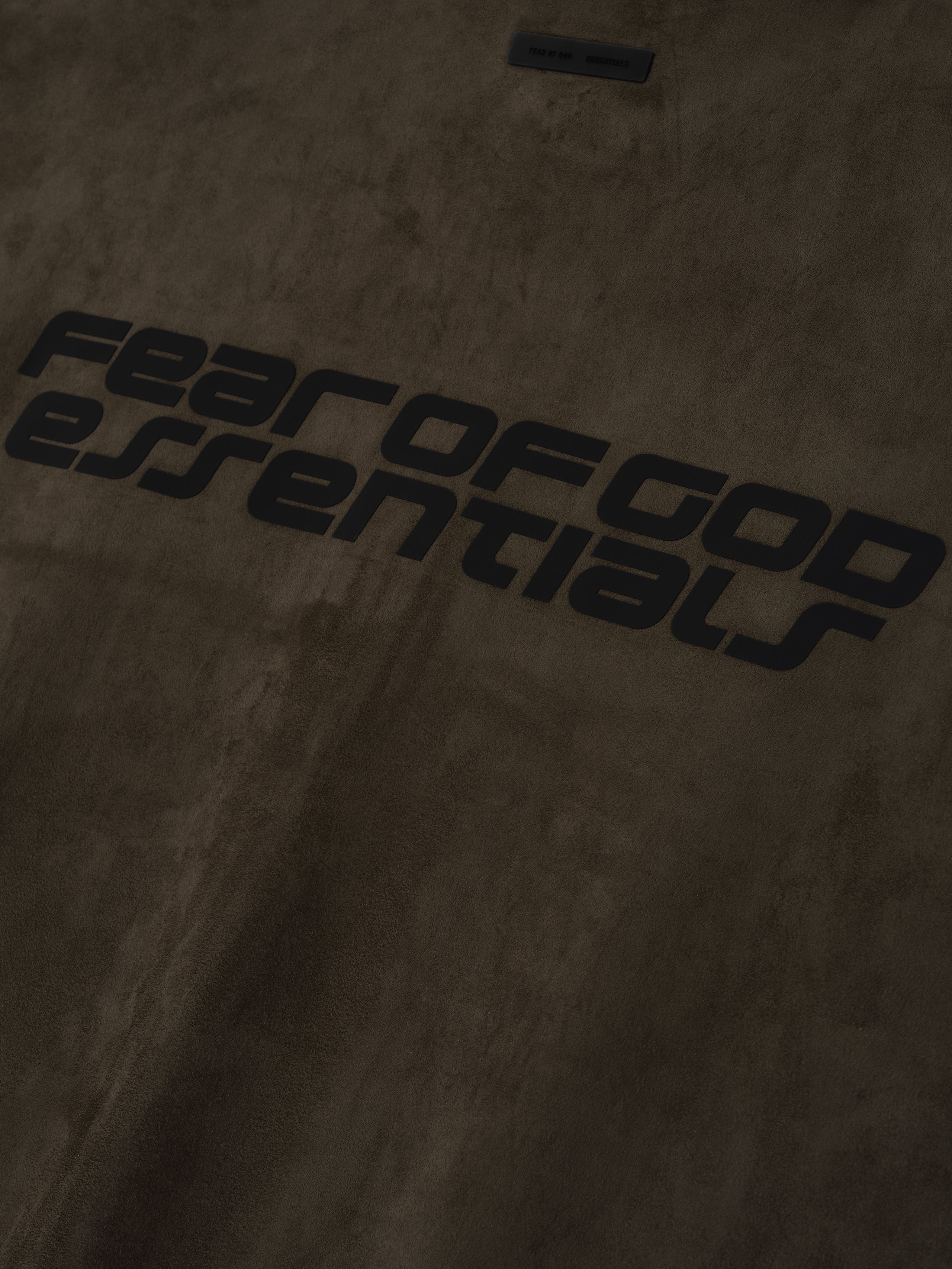 Close-up of brown suede fleece fabric with black Fear of God Essentials logo detailing, file 192HO256952F_HIGH_NECK_3QUARTER_SLEEVE_JACKET-BROWN_3.jpg
