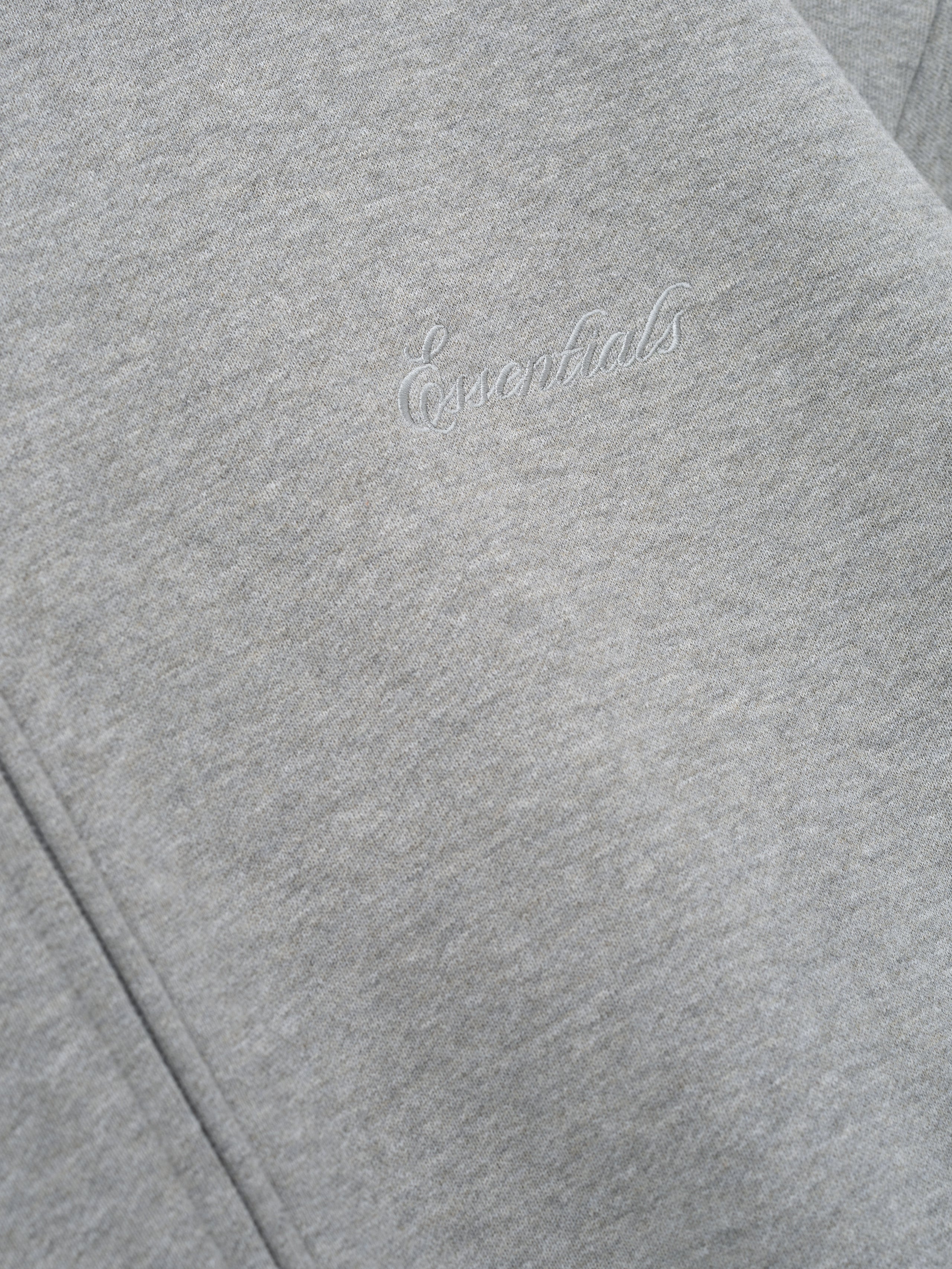 close-up of light gray core fleece hoodie with stitched Essentials signature logo on front panel near zipper