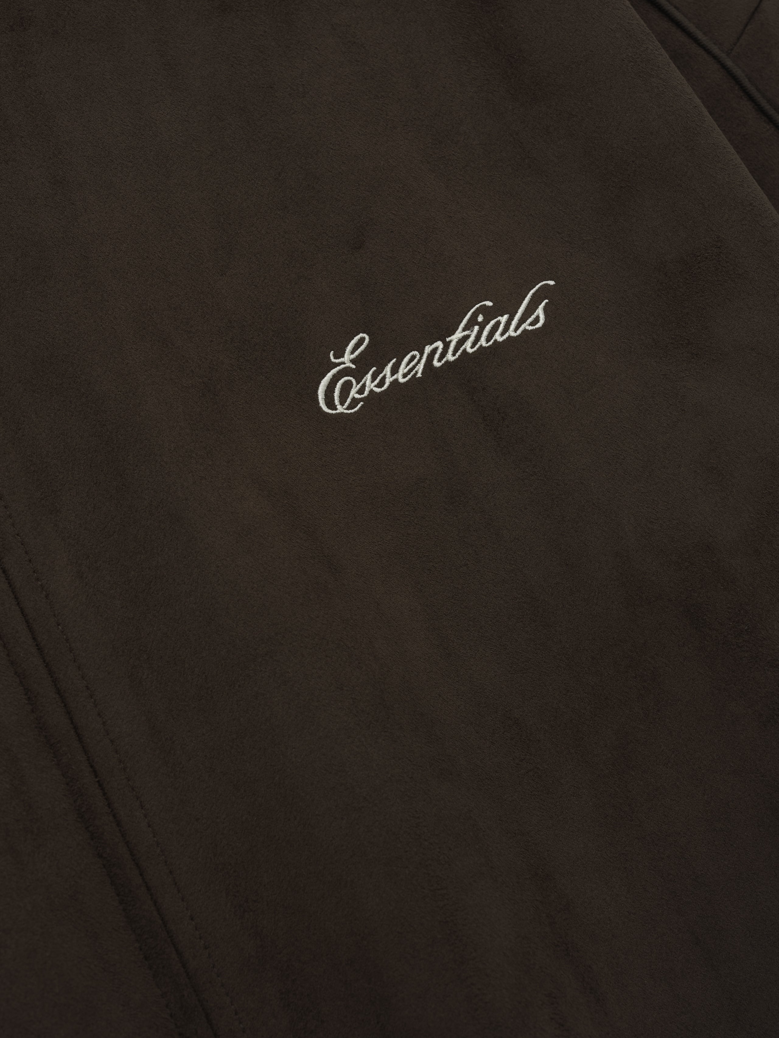 Close-up of dark brown fabric with white cursive embroidered "Essentials" text and visible seam stitching detail