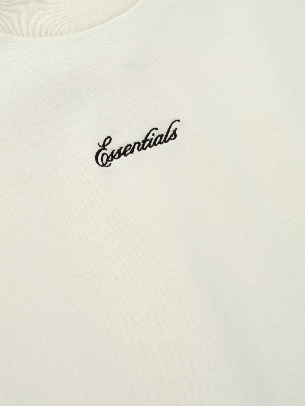 Close-up of the Kids Signature 90s Tee in a soft ivory fabric with black cursive Essentials embroidery on the chest, highlighting a simple and classic kids' top.