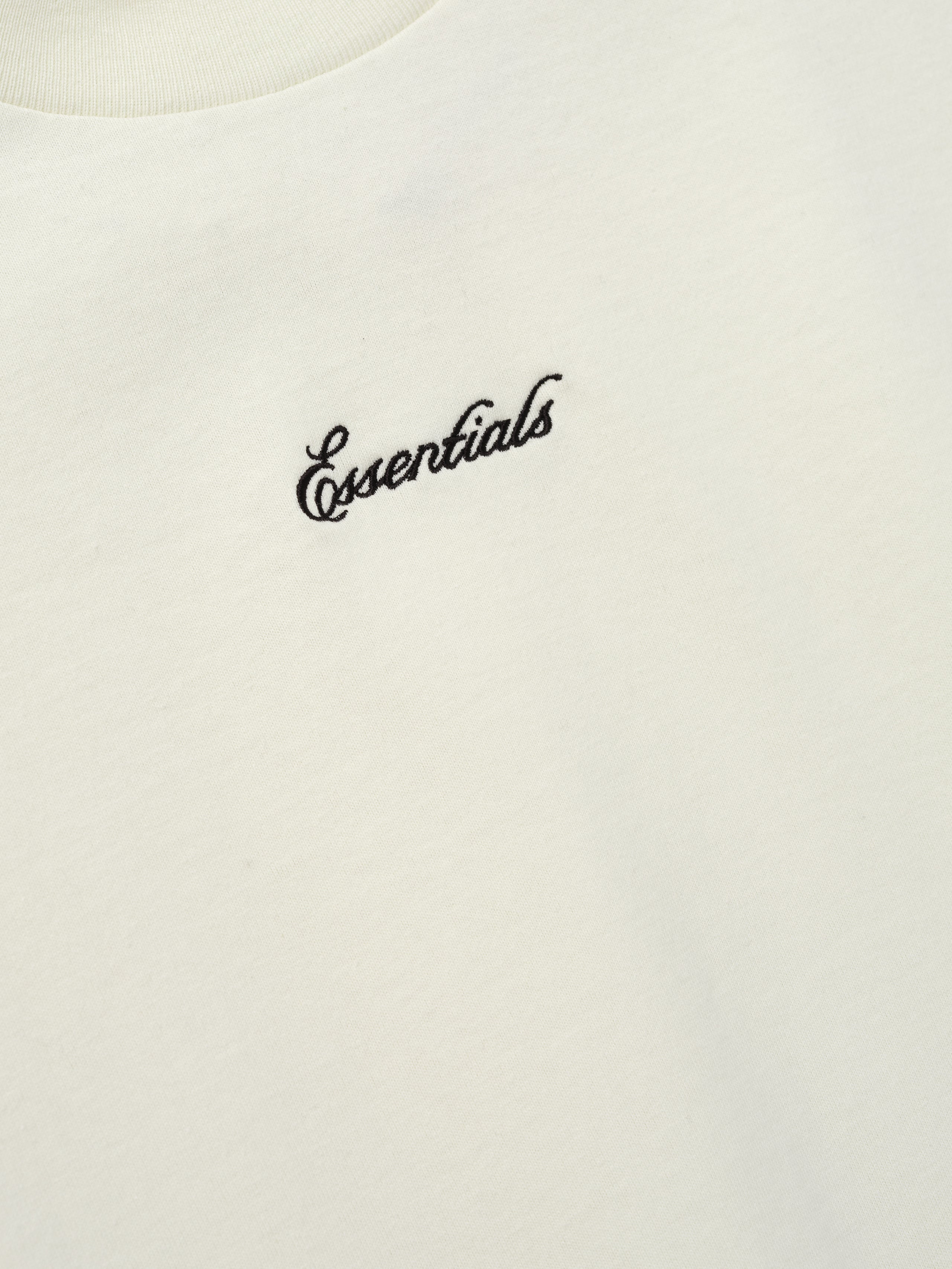 Close-up of the Kids Signature 90s Tee in a soft ivory fabric with black cursive Essentials embroidery on the chest, highlighting a simple and classic kids' top.
