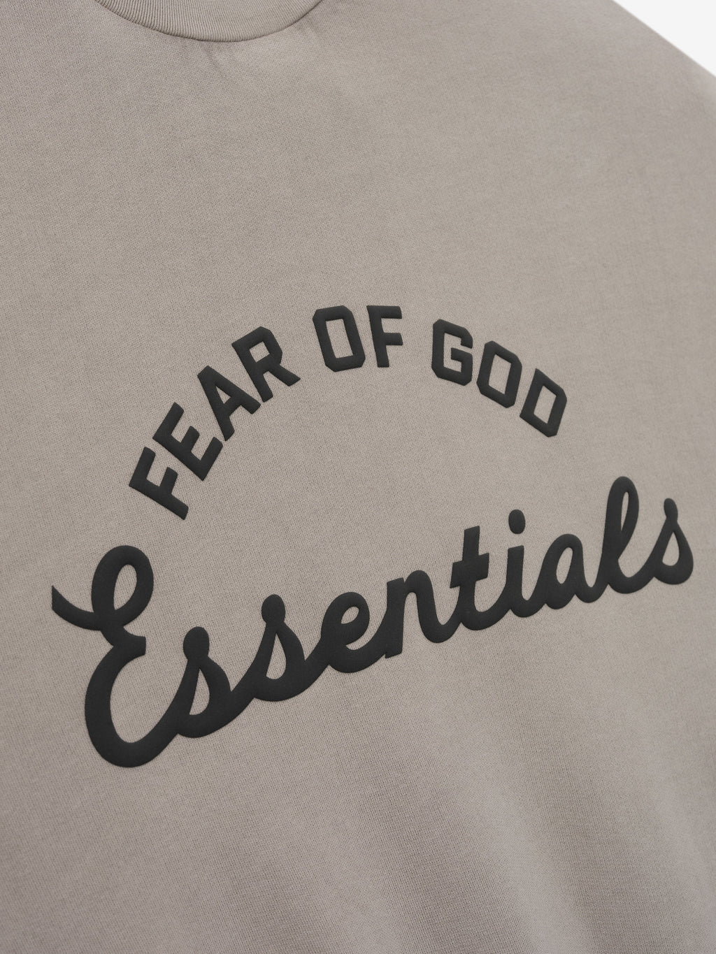 Close-up of taupe fabric with raised black "FEAR OF GOD Essentials" text in arched and cursive fonts on front center