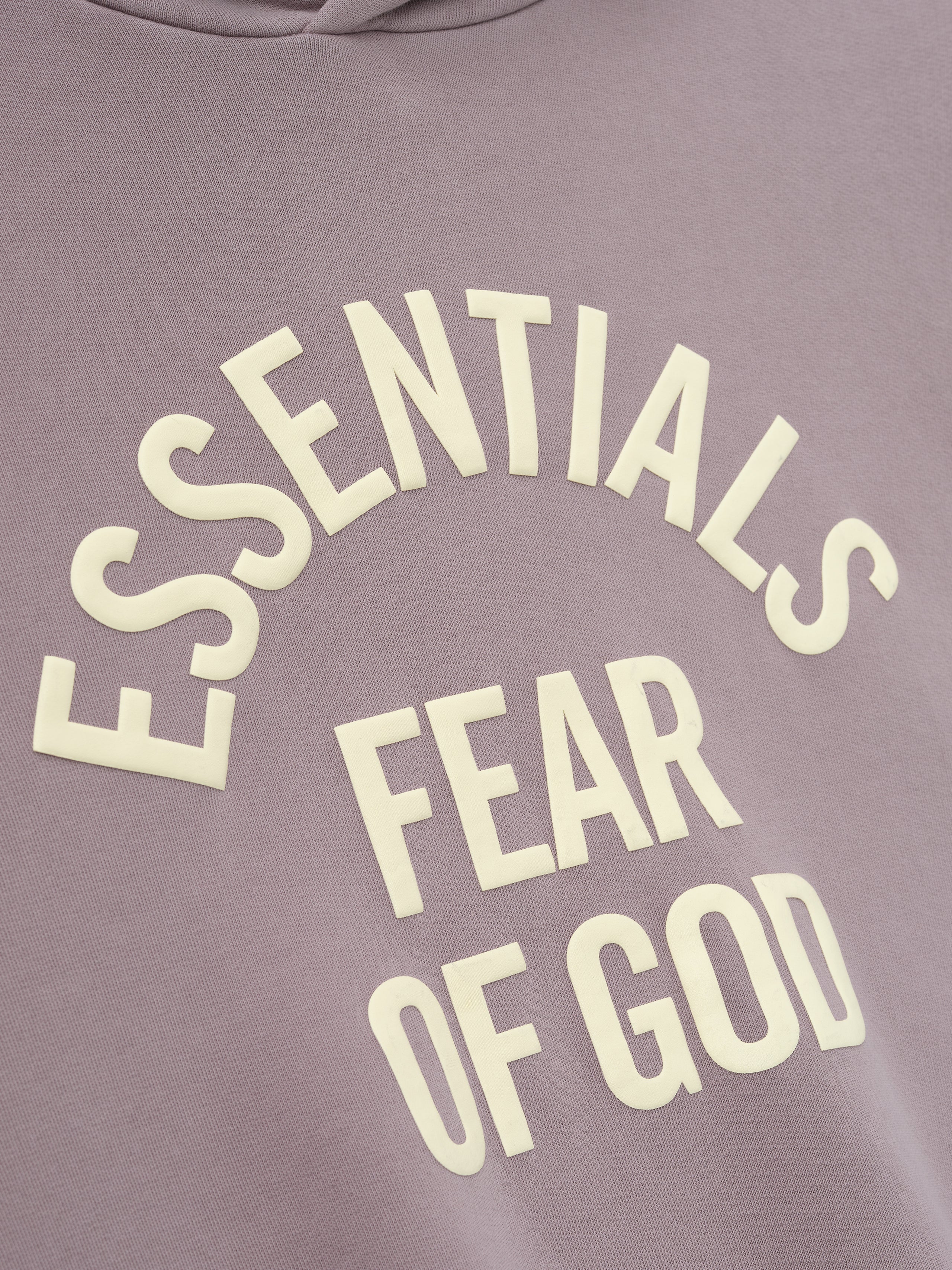 close-up of muted mauve pigment-dyed fleece fabric with raised cream Essentials Fear of God text printed in curved layout
