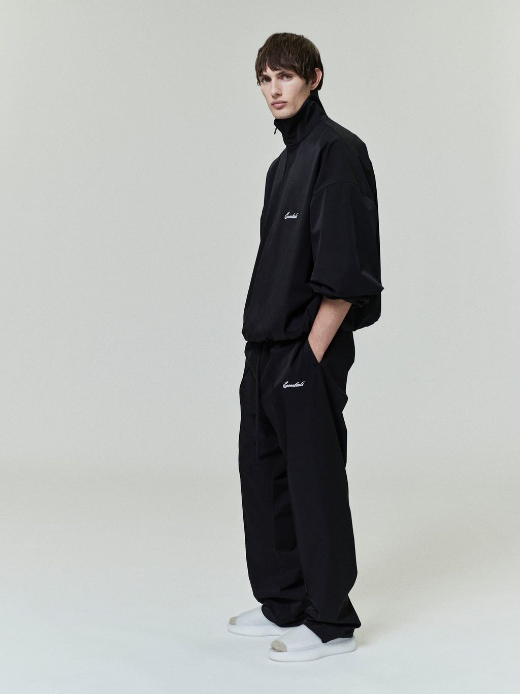 Man wearing a black sporty nylon half-zip pullover and matching relaxed pants with white sneakers on a plain background
