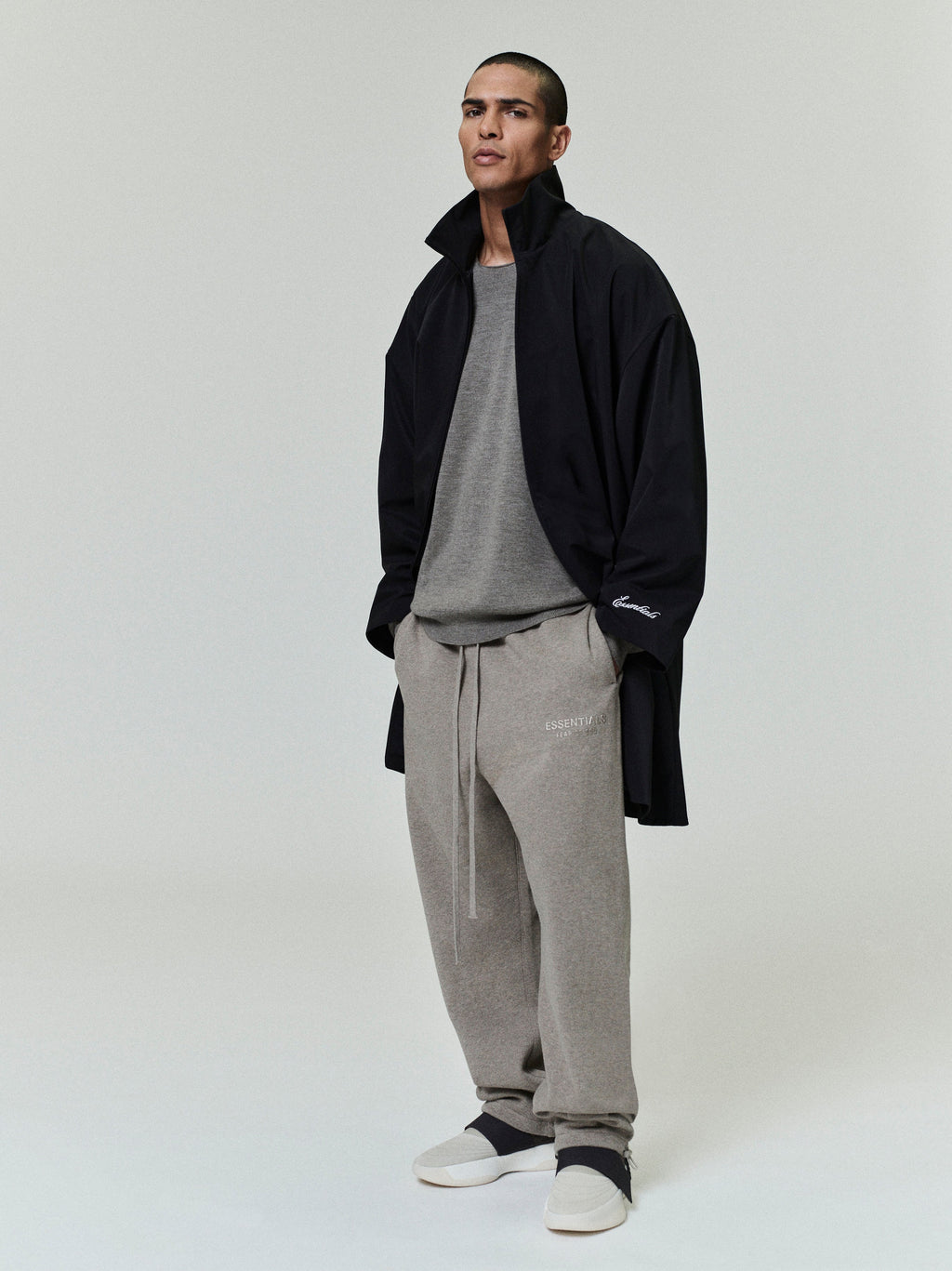 Man wearing a black sporty nylon high neck jacket over a gray shirt and light gray sweatpants with white slip-on shoes