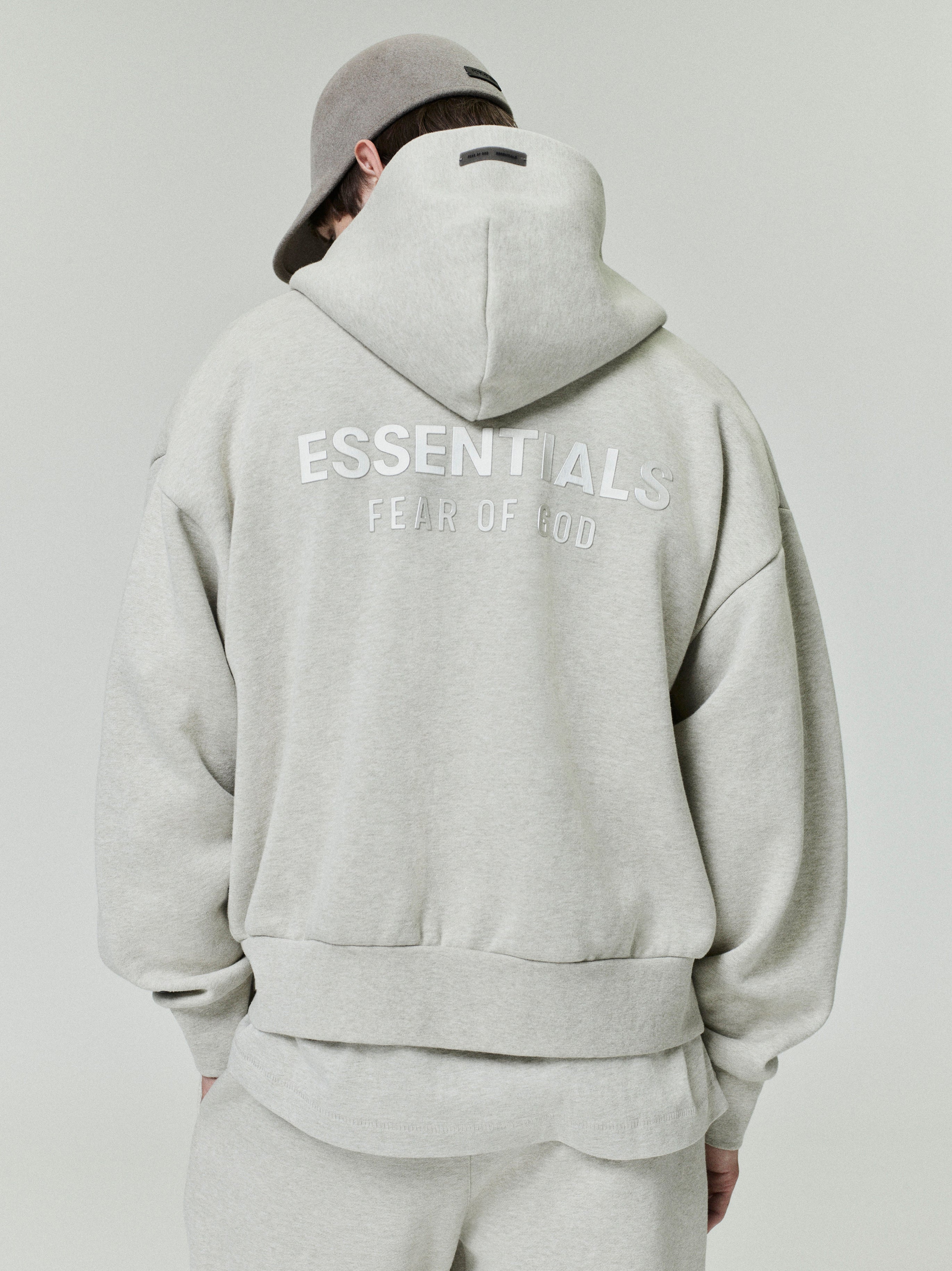back view of light grey oversized fleece hoodie with ESSENTIALS Fear of God logo printed across the upper back