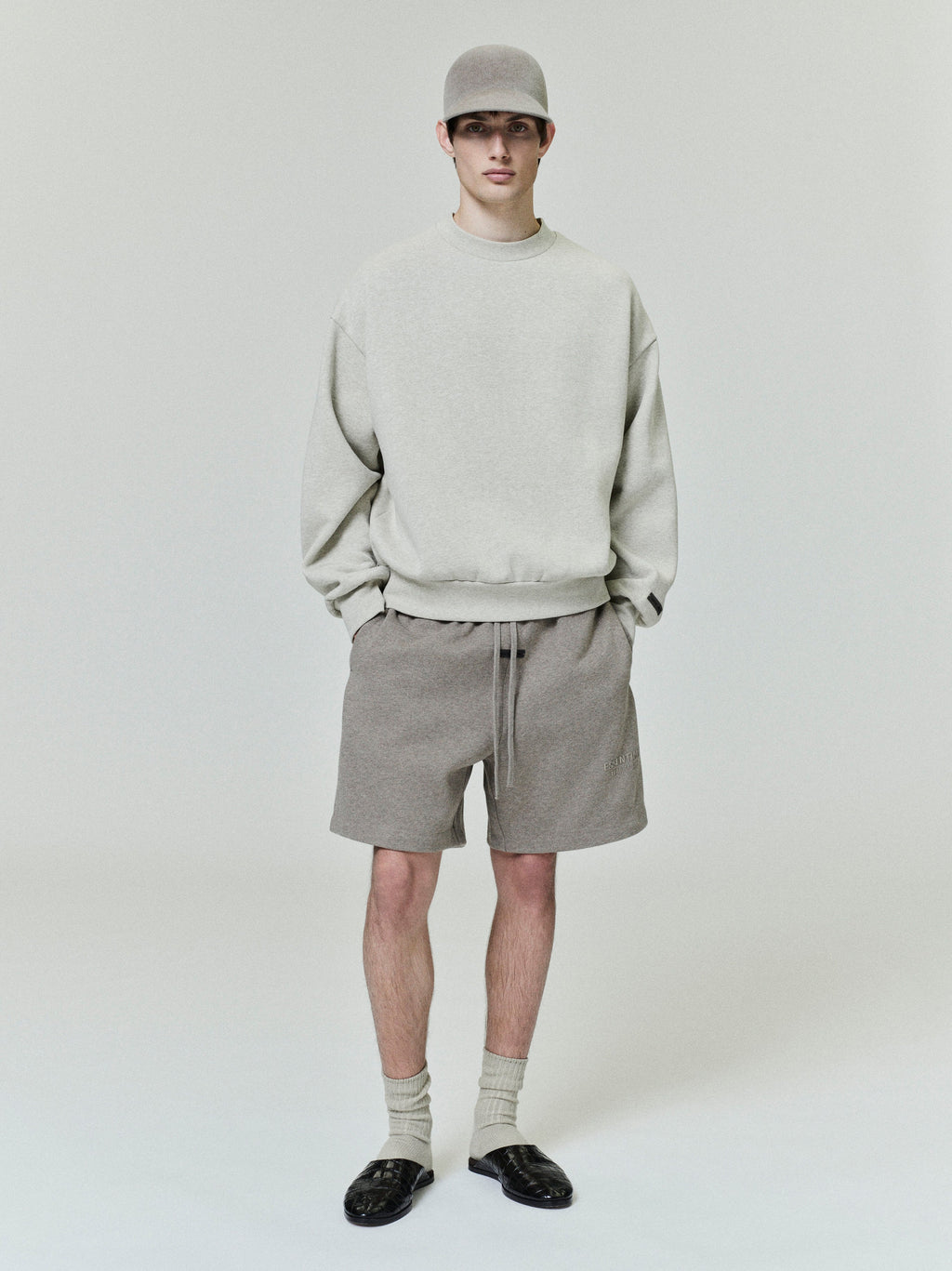 Model wearing oatmeal classic fleece crewneck paired with taupe shorts, matching cap, and black slip-on shoes on plain background