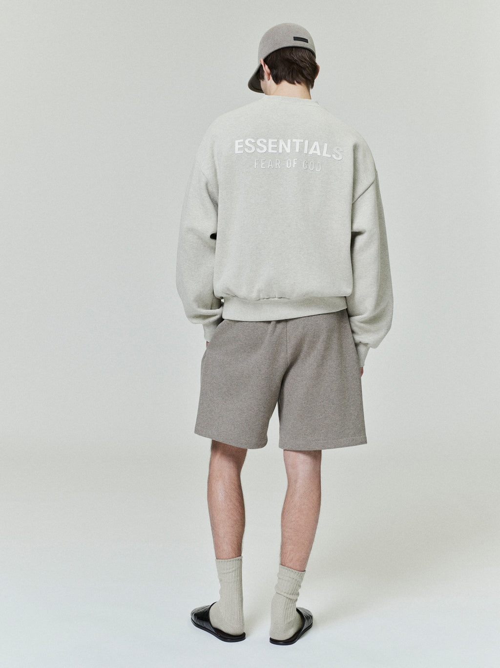 Rear view of model wearing light grey Classic Fleece Crewneck with ESSENTIALS logo paired with taupe shorts and matching cap