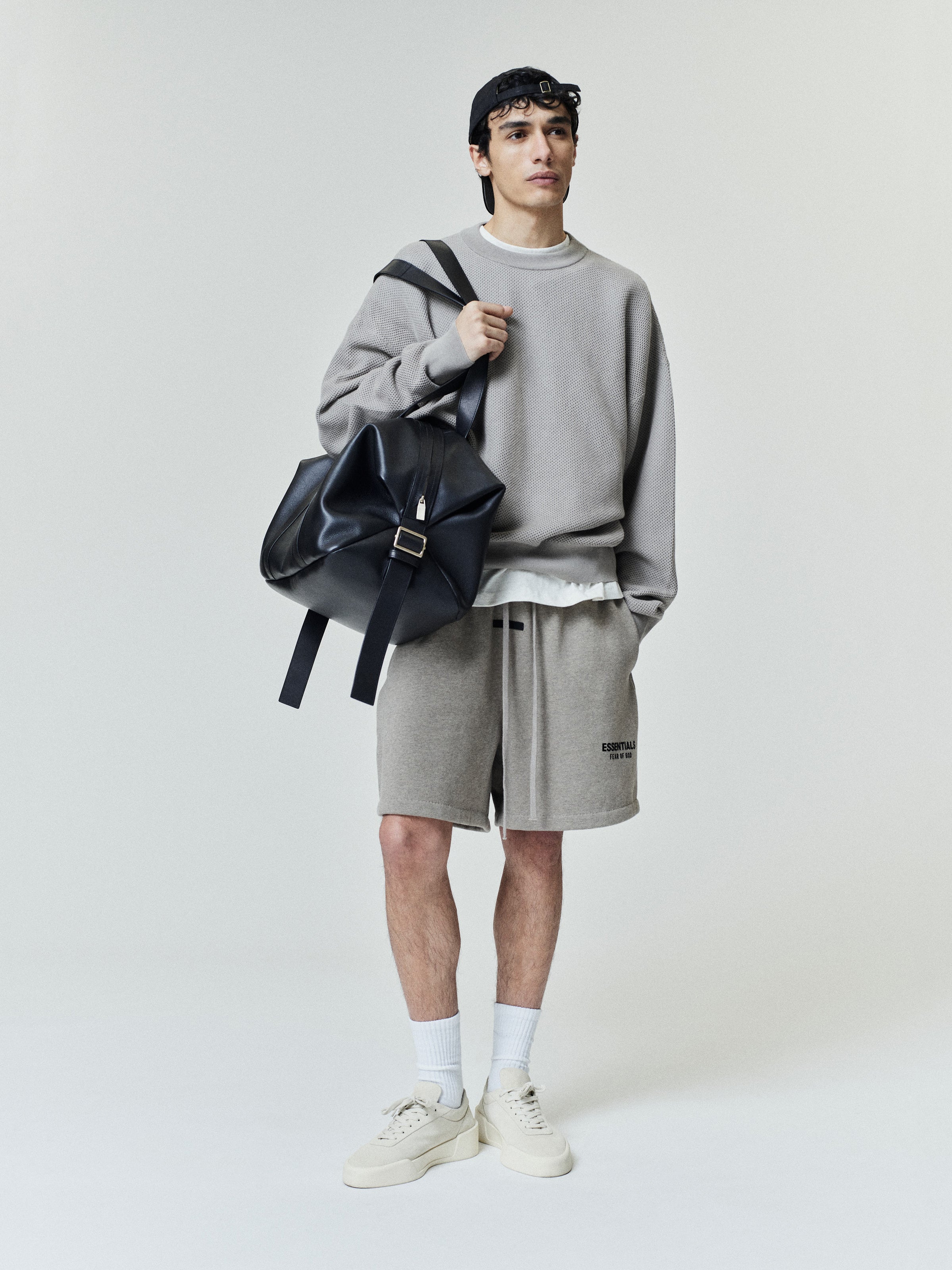 Young man wearing a loose beige sweatshirt over a white shirt, paired with matching knee-length shorts featuring small text on the left leg, white socks, beige sneakers, black cap, and carrying a slouchy black leather shoulder bag.