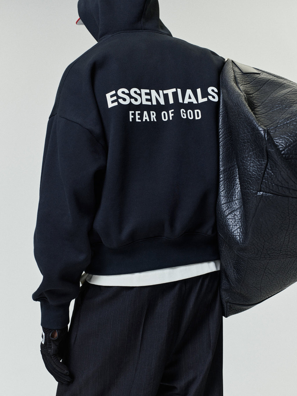 Person wearing a black hoodie with white "ESSENTIALS FEAR OF GOD" text on the back, paired with black pants and holding a large textured black shoulder bag.