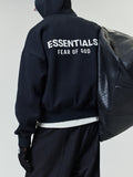 Person wearing a black hoodie with white "ESSENTIALS FEAR OF GOD" text on the back, paired with black pants and holding a large textured black shoulder bag.