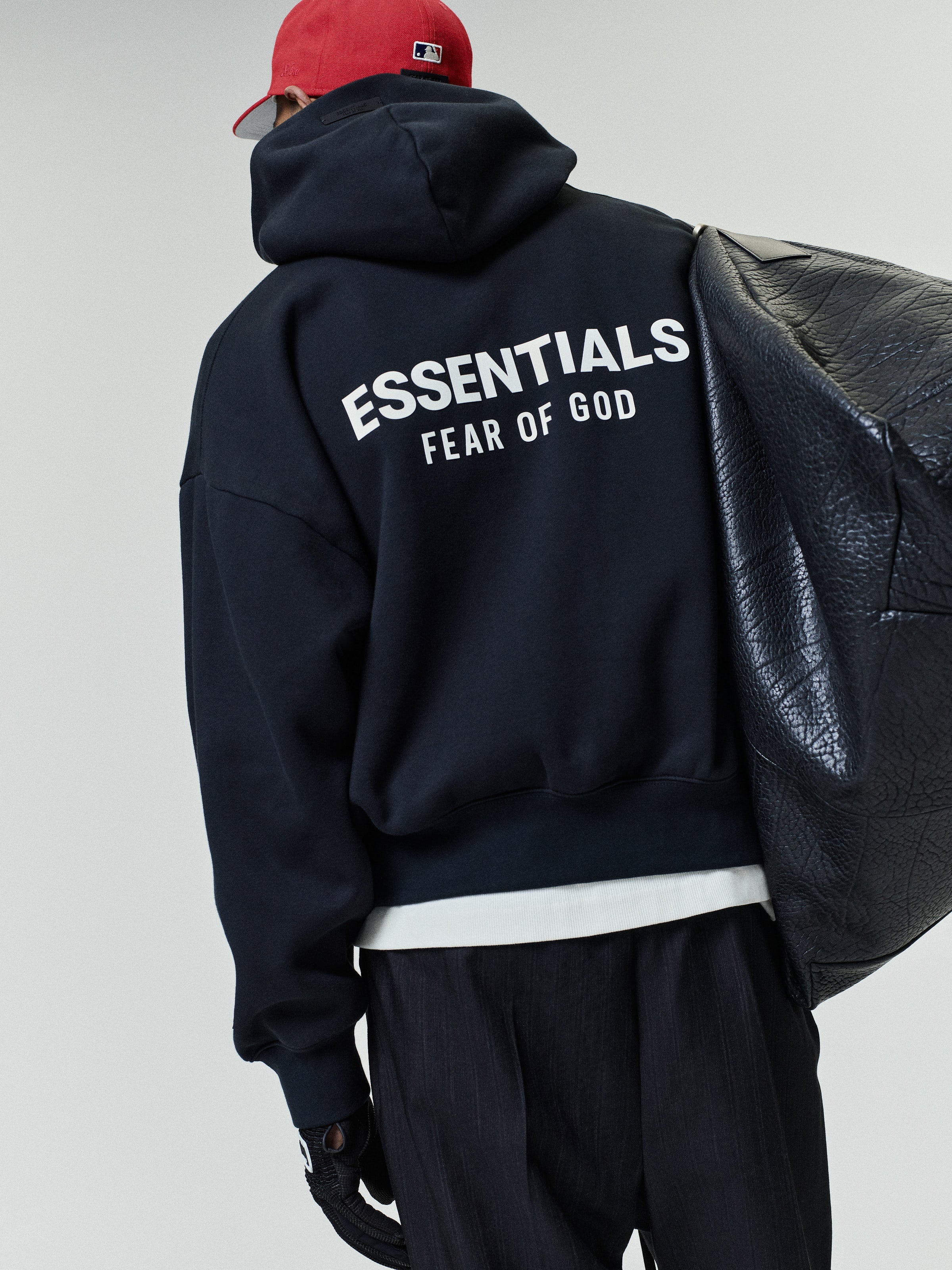 Person wearing a black oversized hoodie with large white "ESSENTIALS FEAR OF GOD" text on the back, paired with black pants, a red cap, and carrying a textured black leather bag.
