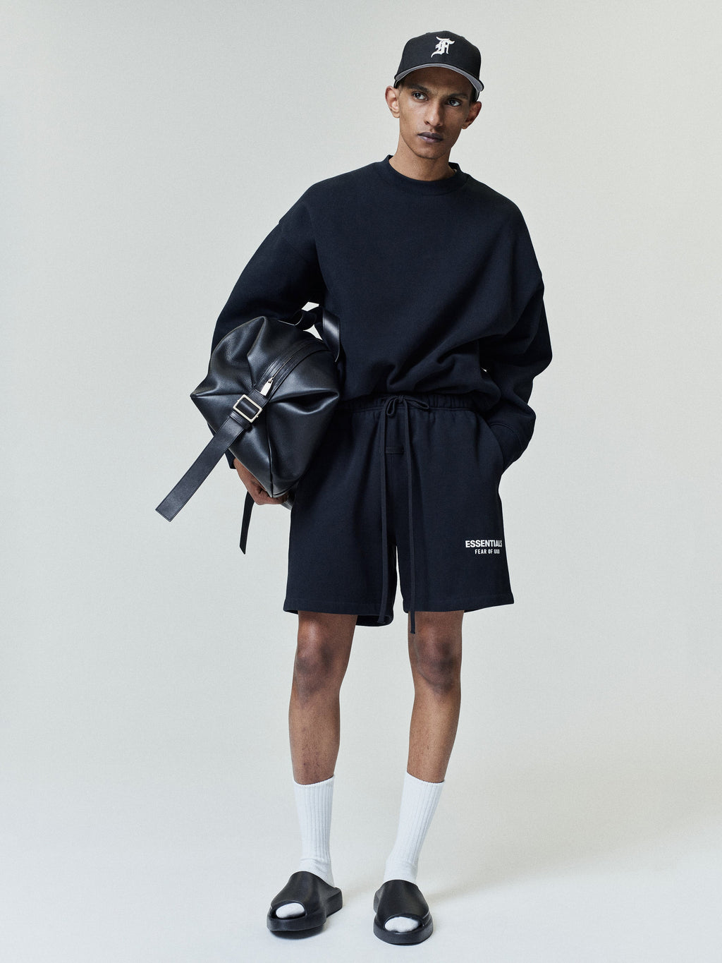 Young man dressed in a black oversized sweatshirt and matching shorts with "ESSENTIALS" text on the left leg, paired with white crew socks, black slide sandals, and a black cap while holding a large black leather bag.